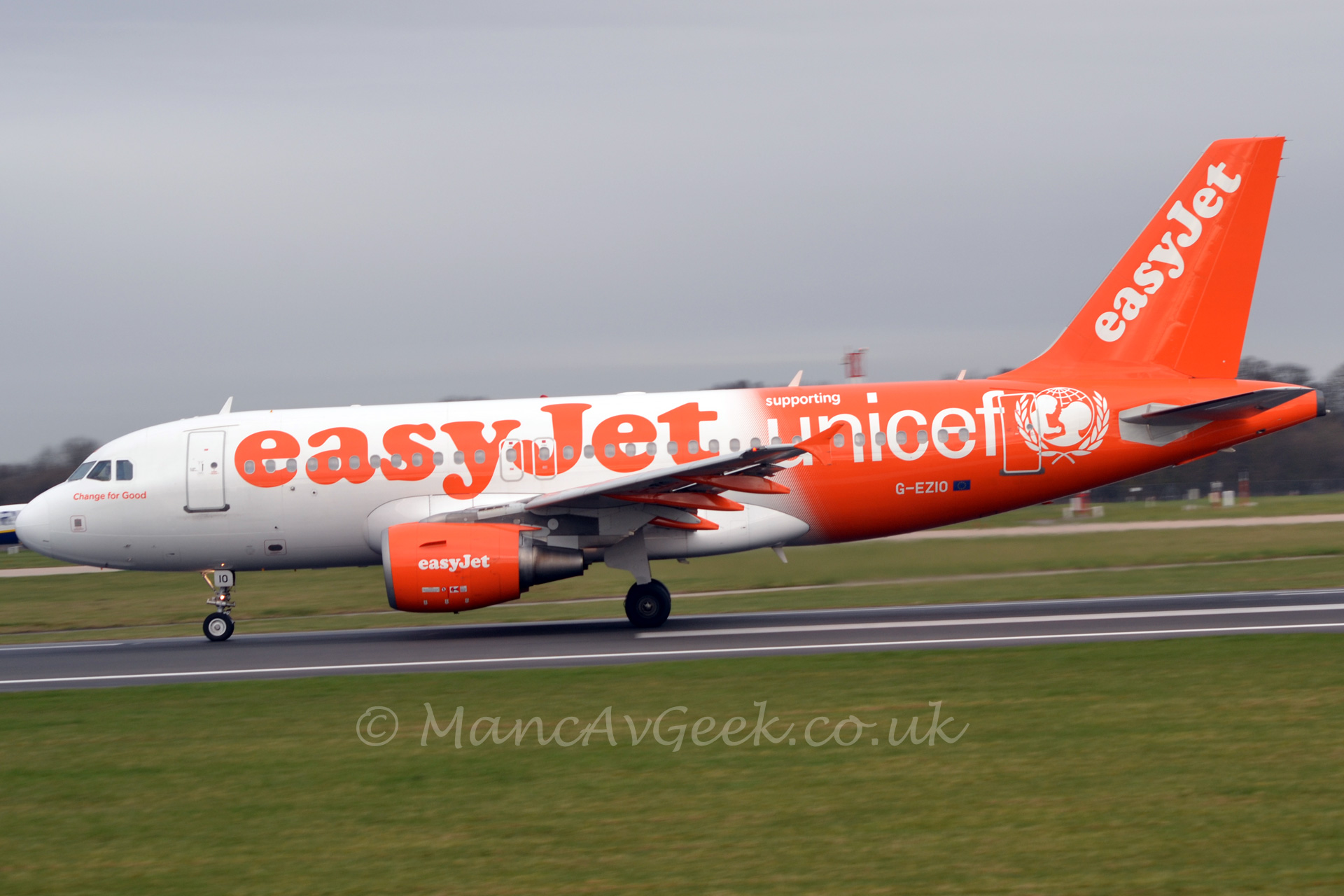 Side view of a twin engined jet airliner accelerating along a runway on it's take-off run. The plane is mostly white, with large orange "easyJet" billboard style titles on the forward fuselage, with smaller orange "Change for Good" titles under the cockpit windows. The rear fuselage is almost entirely orange, with large "supporting Unicef" titles and the unicef logos (an image of a wireframe globe overlaid with an image of the heads of an adult and a child, surrounded by a laurel wreath). The tail and engines are also orange, with white "easyJet" titles. The foreground is mostly grass, while the background is mostly grass below the plane, with some trees visible above the nose and under the tail. Above, the sky is an ominous grey.