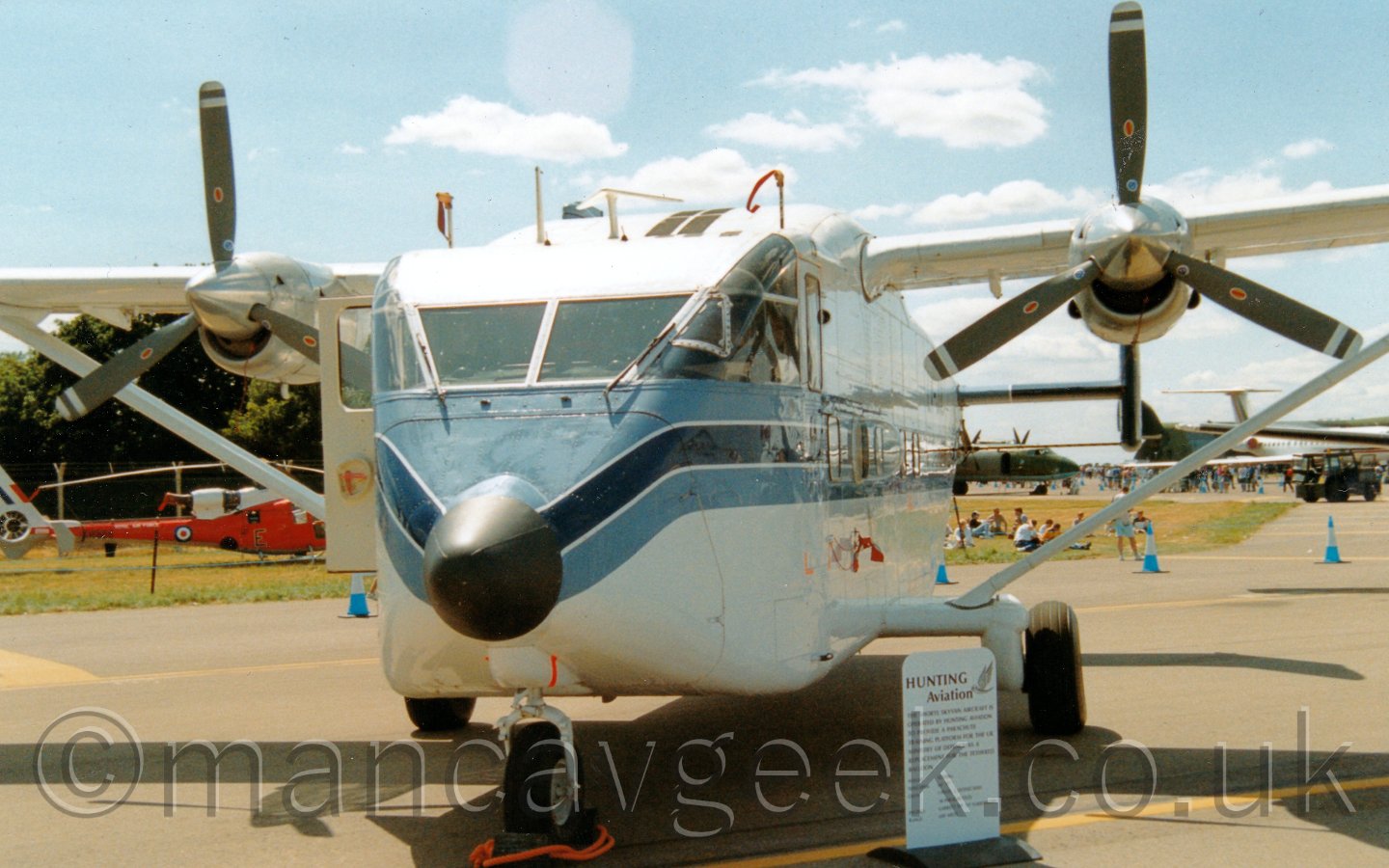 Front view of a high-winged, twin-tailed, twin propellor-engined cargo plane parked facing slightly off to the left of the camera. The plane is mostly white, with a light blue/dark blue/light blue stripe running along the body from the nose. There is a small sign giving a description of the plane on the ground by the nose in front of the left main wheel. In the background, a red helicopter is visible in front of trees on the left of the frame, while several green or white transport aircraft are visible on the right. Above and behind it all the sky is a gorgeous blue, with lots of fluffy white clouds here and there.