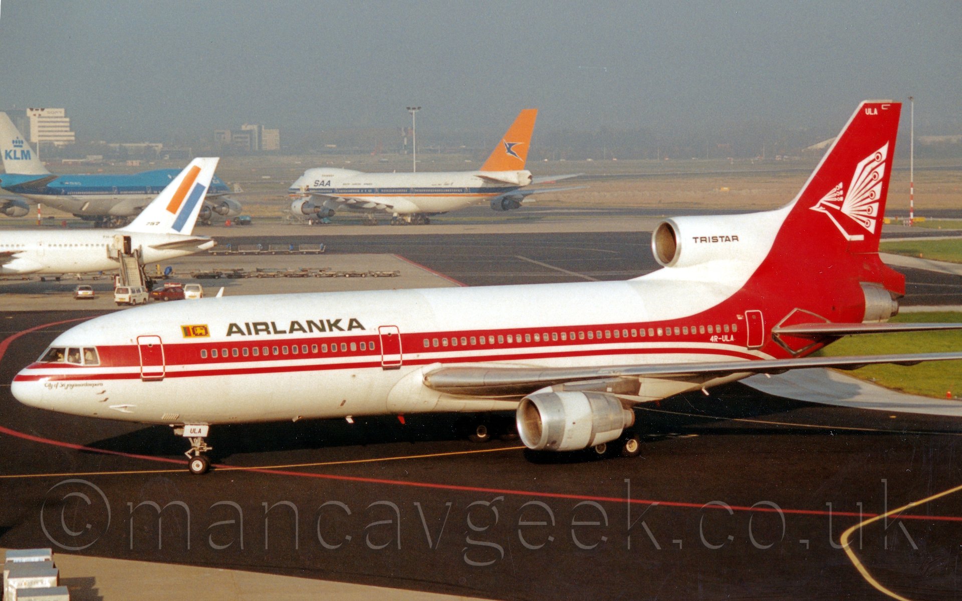 Side view of a 3 engined jet airliner taxiing from right to left. The plane is mostly white with a thick red line running rearward from the nose, covering the cabin windows, before sweeping up to encompass most of the tail and rear fuselage. There are "Air Lanka" titles on the upper forward fuselage, and a white stylised flying bird on the tail, with additional "TRISTAR" titles on the centre engine air intake In the background, there are seveeral aircraft parked, with large grassed areas leading off into the distance, eventually vanishing in to haze.