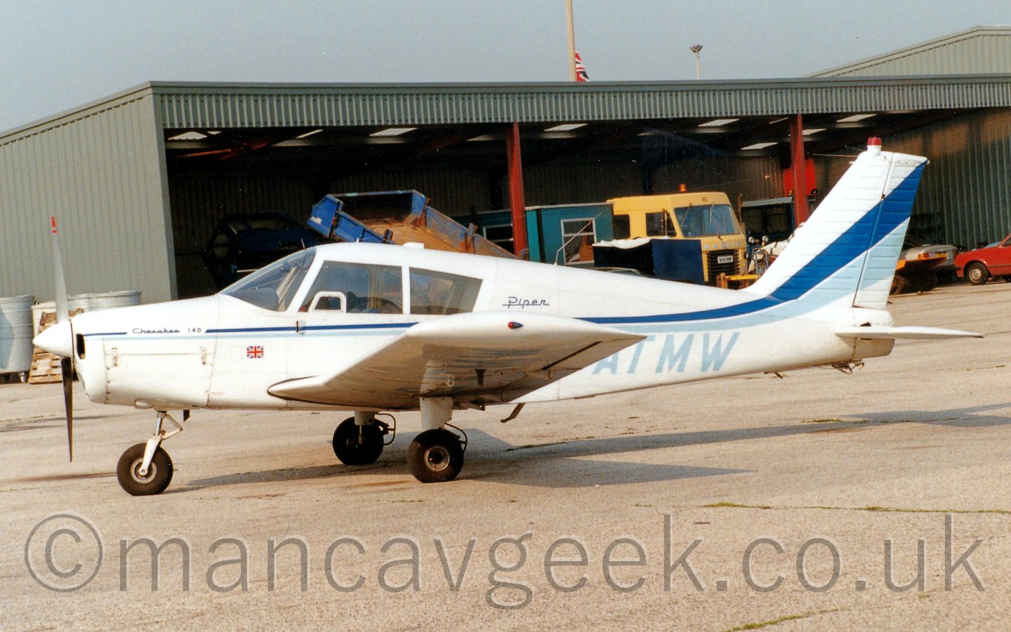 Side view of a single engined light aircraft parked facing to the left. The plane is mostly white, with a light and dark blue stripe runnning along the fuselage and sweeping up in to the tail.behind is a large grey metal shed with several vehicles and a cabin parked inside. The sky above is a light grey.