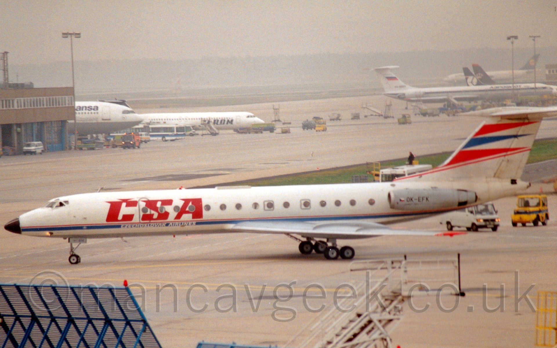 Side view of a twin engined jet airliner with a T-tail and the engines mounted on the rear fuselage, taxiing from right to left. The plane is mostly white, with a thin red and blue line runnning along the body, with large "CSA" titles on the forward fuselage, with smaller "Czechslovak Airlines" underneath. The tail is white, with what appear to be red, blu, and red pennants pointing backwards. There are a couple of airport vehicles behind and below the tail, while in the background, several parked planes vanish into grey haze in the distance.