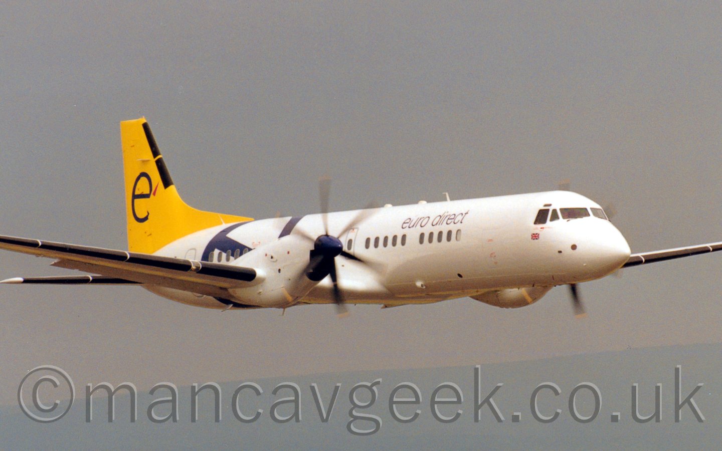 Side view of a twin propellor airliner flying al low level from left to right. The plane is mostly white, with a large blue letter "e" on the rear fuselage, and "euro direct" titles on the upper forward fuselage. The tail is all yellow, with another blue "e" in the middle. Behind, the sky is a dreary grey.