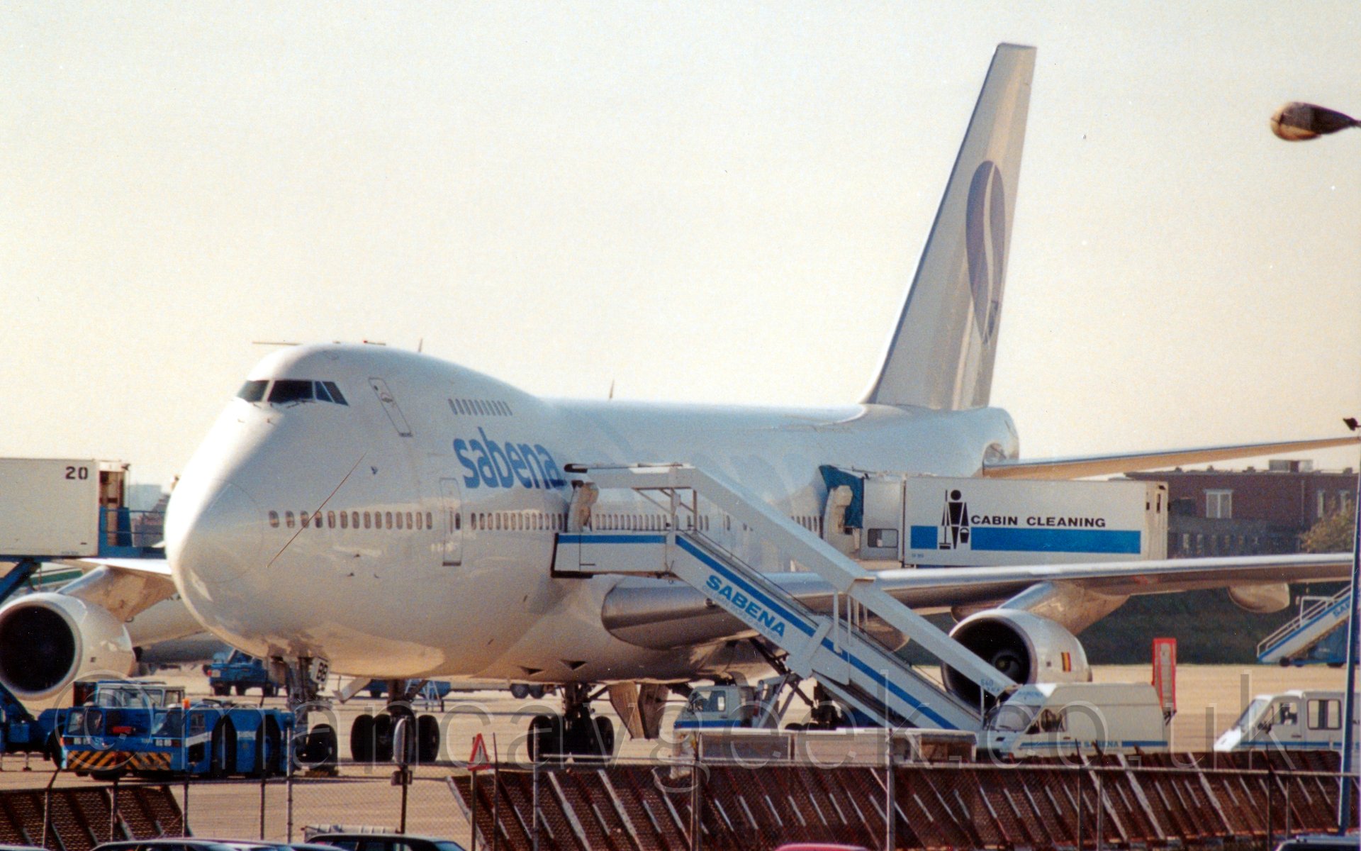 Front view of a very large 4 engined jet airliner parked facing slightly to the left of the camera. the plane is mostly white, with light blue "Sabena" titles on the upper forward fuselage, and larger, much paler blue "Sabena" titles along the whole length of the body. There is a set of mobile airstairs mounted on the back of a truck attached to a lower deck door just forward of the wing, while a cabin cleaning truck is attached to a door just aft of the wing. There are various other vehicles scattered around the plane, including a blue tug attaching itself to the nosewheel. Above it all, the sky is just a mass of hazy cloud.