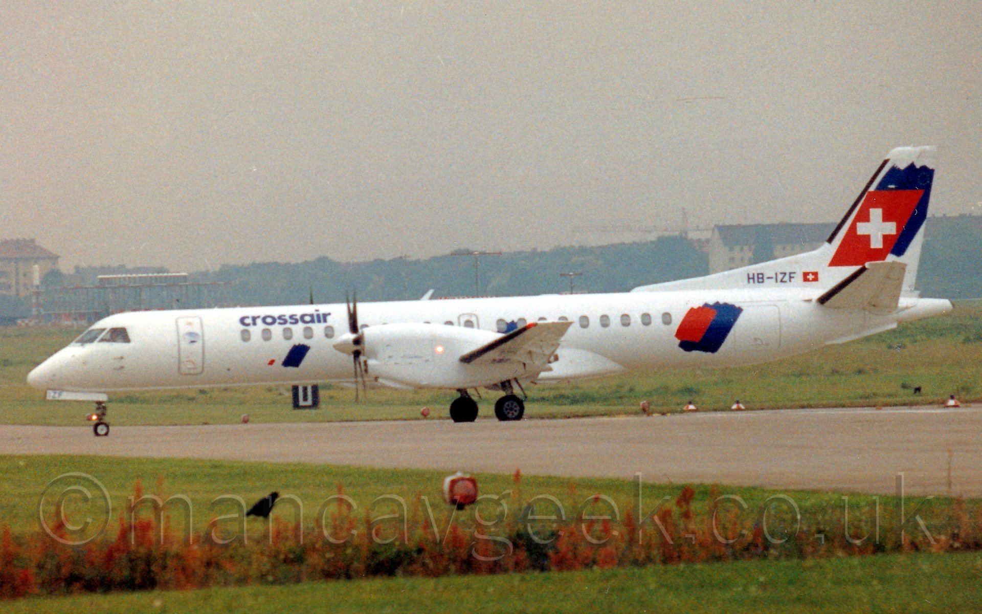 Side view of a twin propellor engined airliner taxiing from right to left. The plane is mostly white, with blue "crossair" titles on the forward fuselage, with red and blue splodges on the rear fuselage. The same splodges are on the tail, the red one being squared off and having a white cross in the middle. The foreground has a large grassed area, with a line of flowering plants, while the background has more grassed areas leding up to tall buildings and trees in the hazy distance. The sky is a hazy grey.