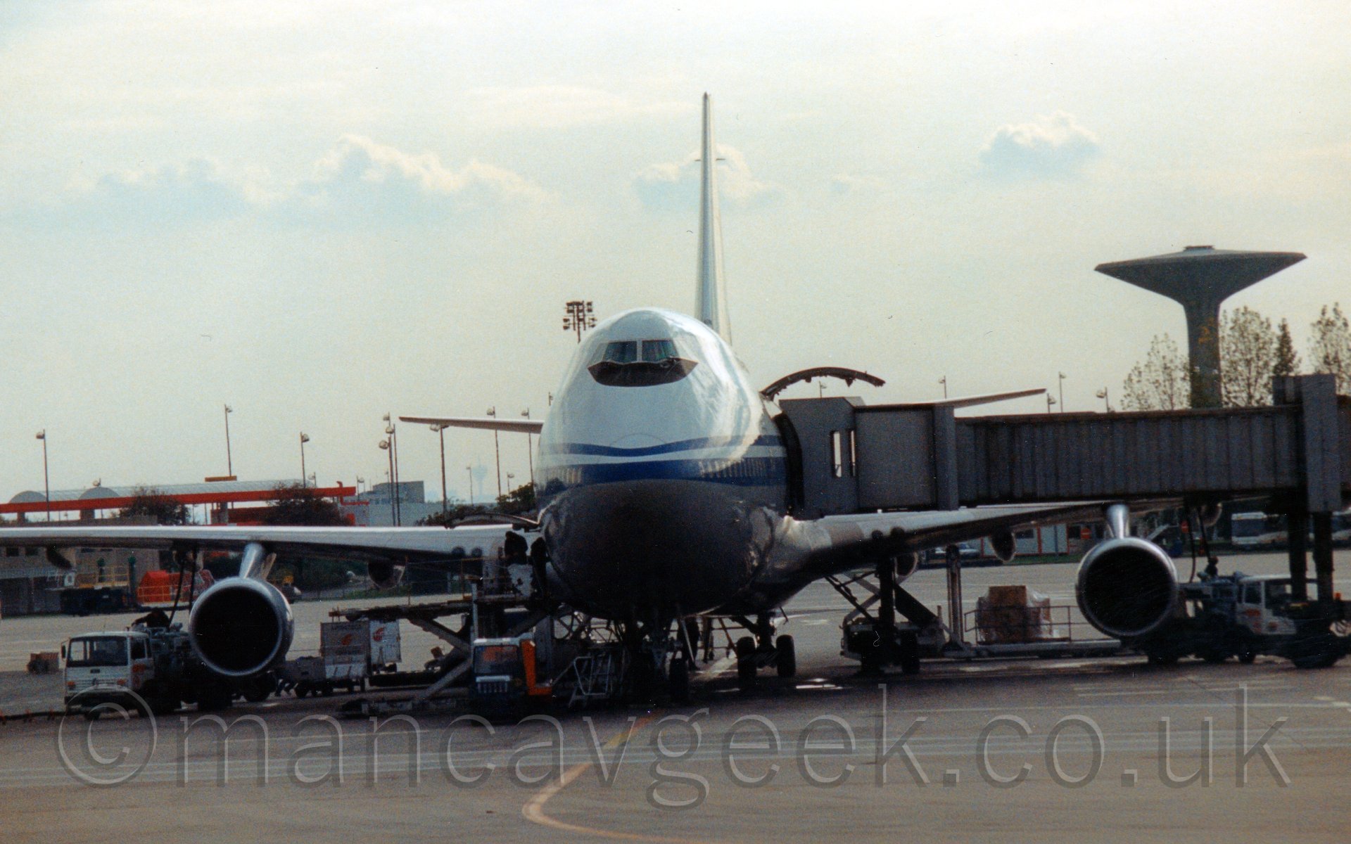Front view of a very large 4 engined jet airliner parked almost head on to the camera, with an air bridge attached to it's left side. A large door in the side of the fuselage is open behind the air bridge, with a large fuselage panel appearing to hang over the bridge. The plane is mostly white, with a grey belly, and a blue stripe running back along the fuselage from the nose. There are various fuel tankers and baggage carts around the eingines and wheels of this plane. The planes tail reaches up into the sky, which is a flat white with occasional lower fluffy clouds.