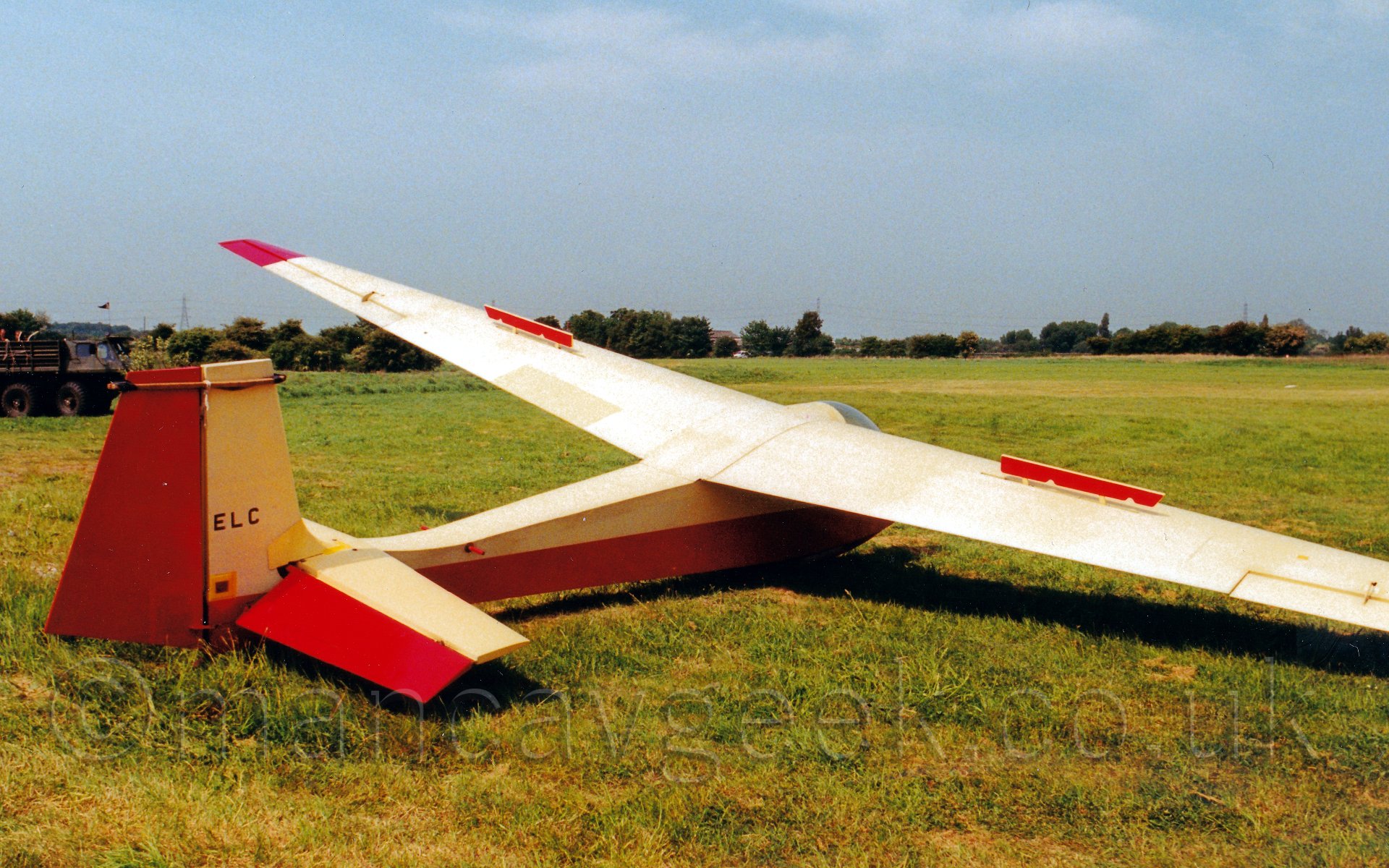Side view of a single seat glider parked on grass, facing to the right and away from the camera, with one wing resting on the ground, the other up in the air. The glider has a red belly, with creamey-yellow upper surfaces, with the rear half of the tail is also red. In the background, the grass airfield stretches into the distance, leading up to lines of trees on the perimeter. Above, the sky is a hazy blue.