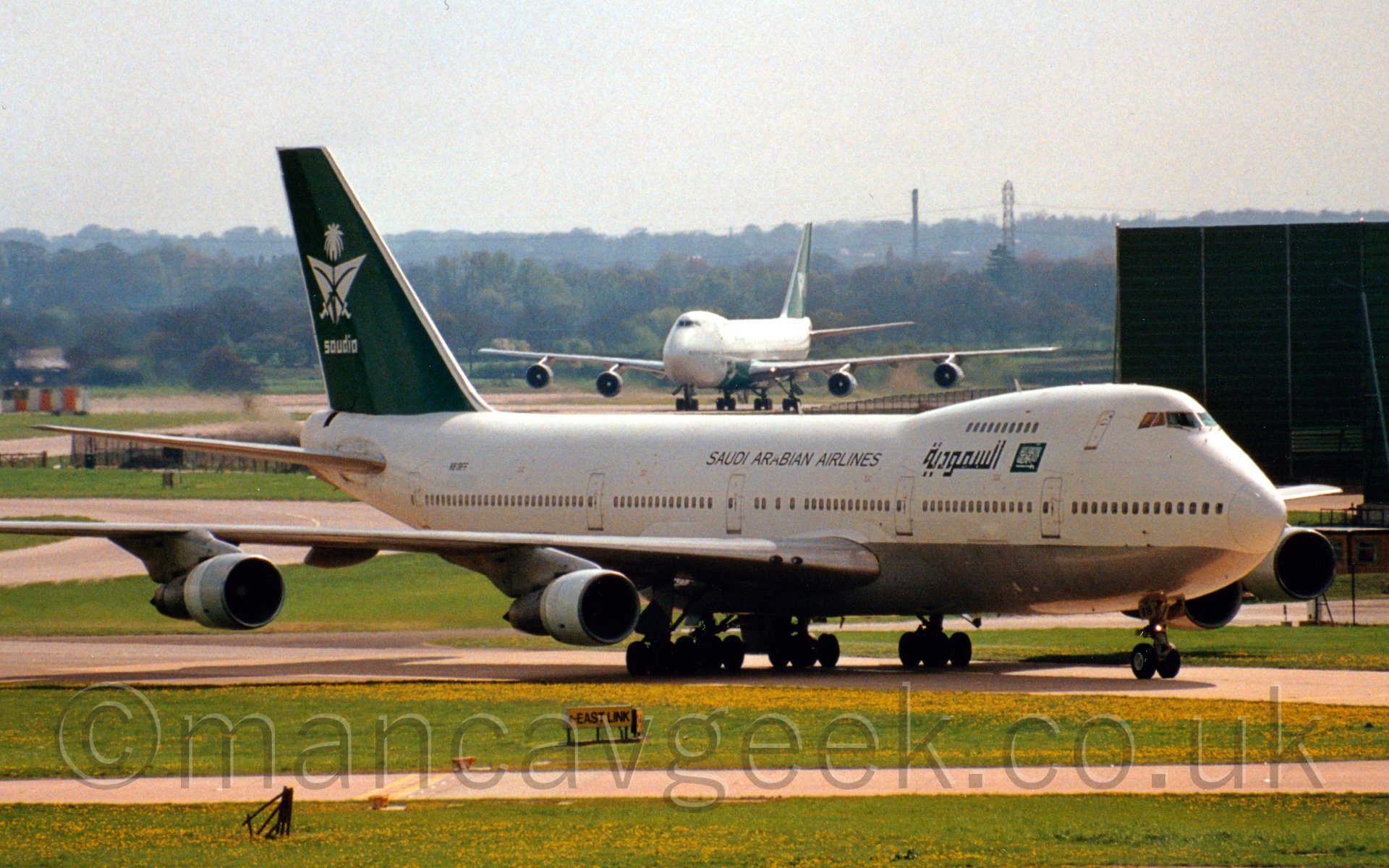 Side view of a very large 4 engined jet airliner taxiing from left to right. The plane is mostly white, with a silver belly, There are black "Saudi Arabian Airlines" titles in English on the upper forward fuselage, starting over the wings, with similar titles further forward in an Arabic script. The tail is a dark green, with crossed curved sword over a palm tree and and inverted chevron logo, with "Saudi" titles underneath. Behind is a similar plane, with trees in the far background, under a misty grey sky.