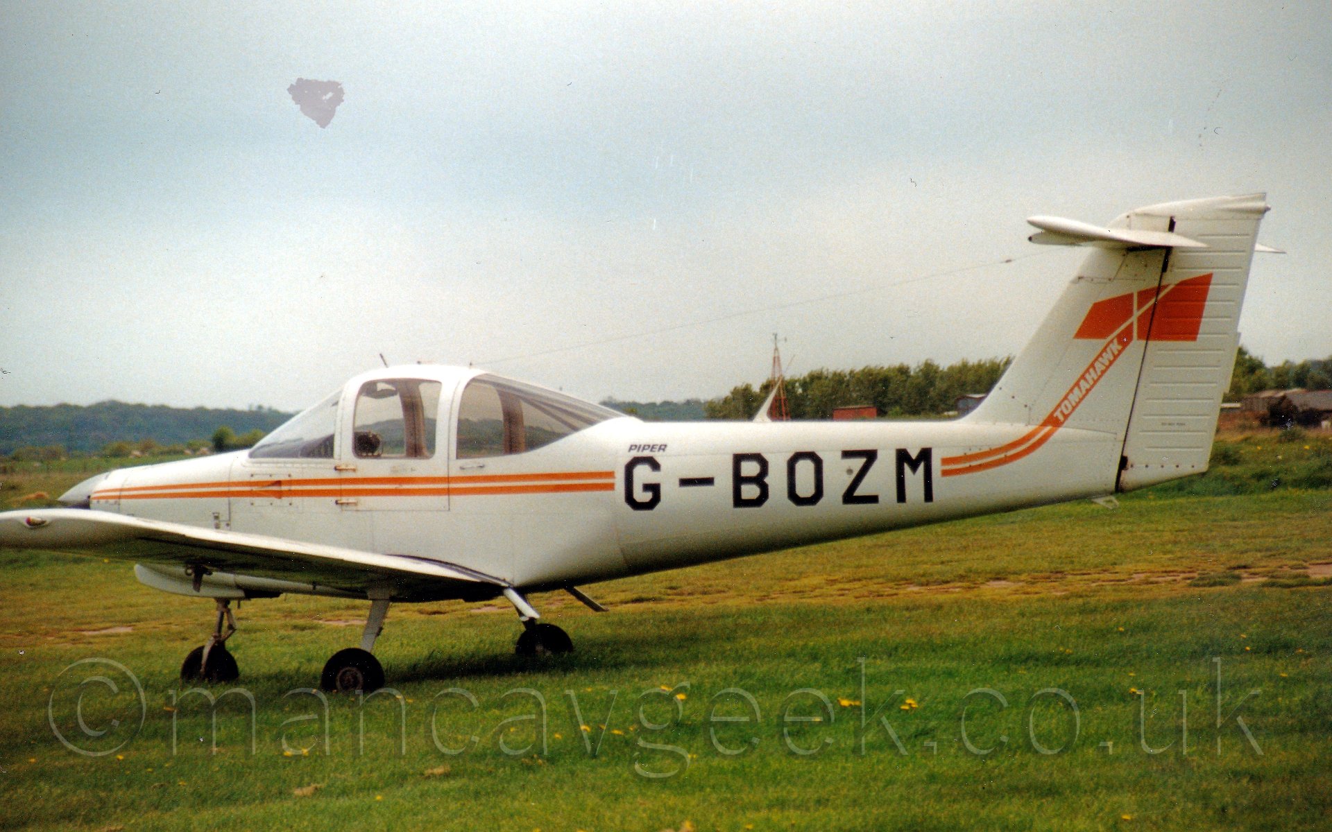 Side view of a single engined light aircraft parked on grass. The plane is mostly white, with a pair of thin orange stripes running along the fuselage, turning into an axe on the tail, with the word "Tomahawk" on the handle in white. The registration "G-BOZM" is displayed prominently on the fuselage. In the background, trees and bushes run to the horizon, under a grey sky.