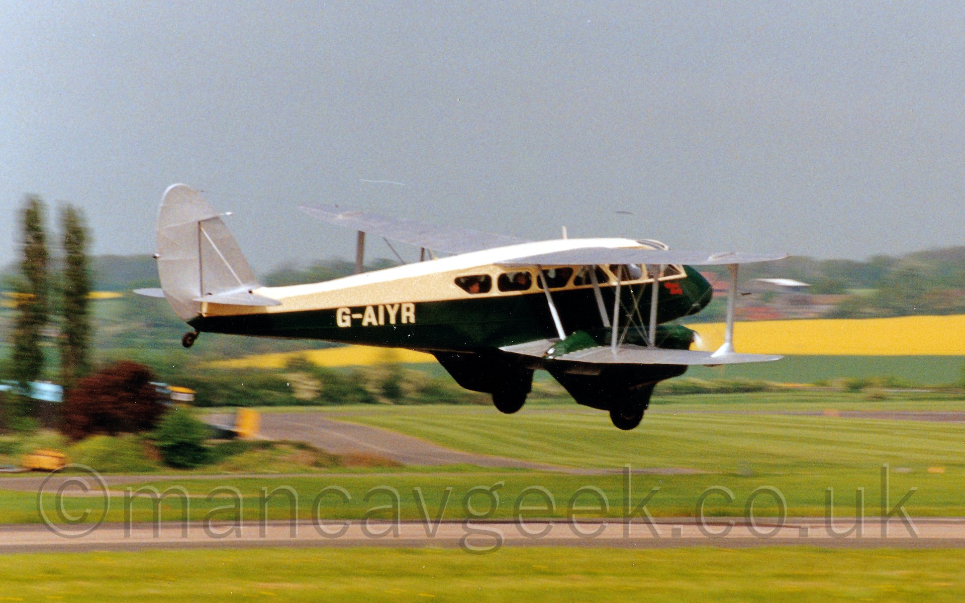 Side view of a twin engined biplane airliner flying at low level from left to right. The plane has a dark green lower half and creamy yellow upper section, with the registration "G-AIYR" also in the same creamy yellow. People are visible through the many large cabin windows.The engines mounted on the lower wings are the same green as the fuselage. In the background, large grassed areas lead off to yellow and green fields, surrounded by large areas of trees., all under a blue-grey sky.
