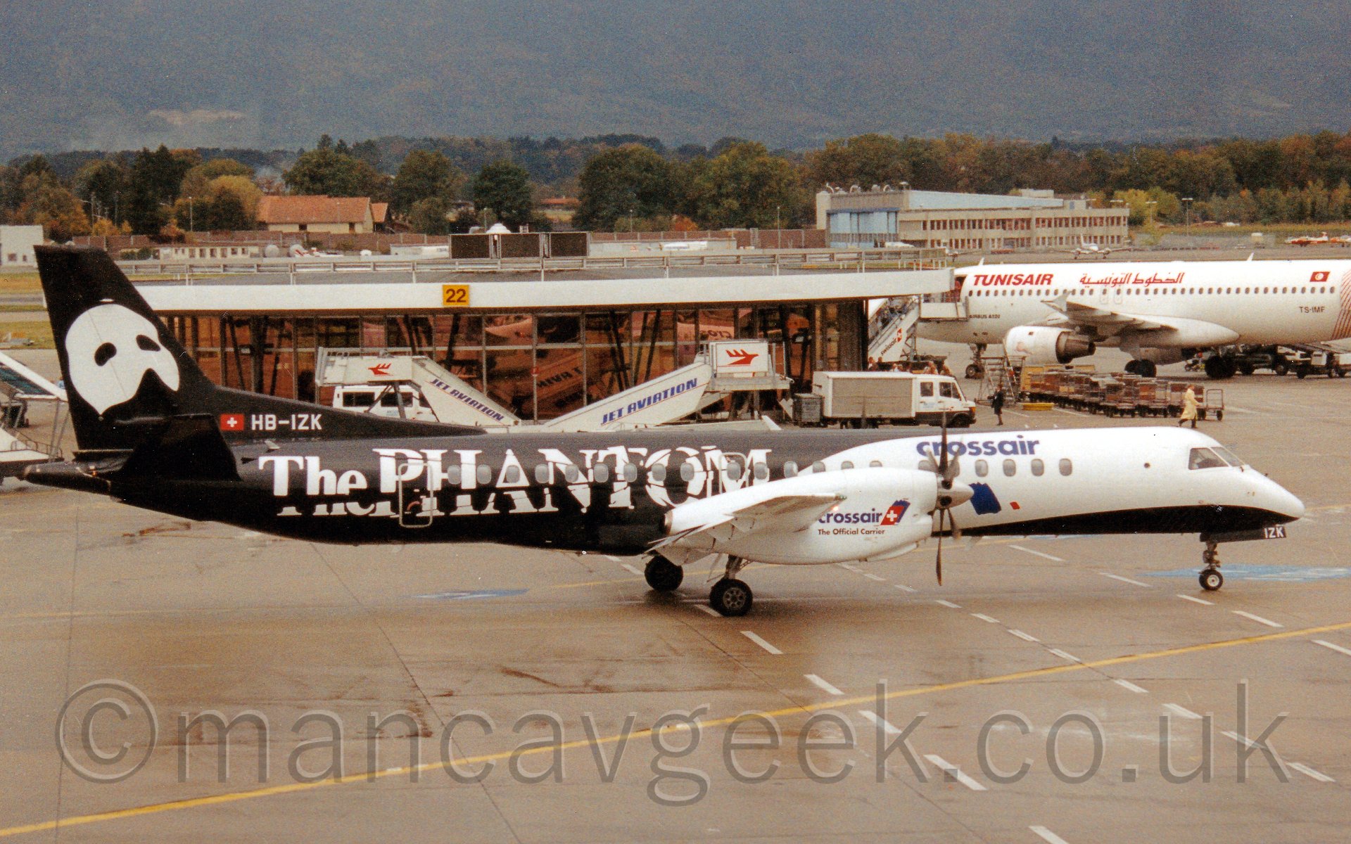 Side view of a twin propellor engined air liner taxiing from left to right. The plane is mostly black, with a large white area on the forward fuselage, with blue "Crossair" titles over the cabin windows towards the front. The rear fuselage has white "The Phantom" titles in a fractured font, while the tail has a white half-face mask. The wing-mounted engines are white, with "crossair" titles. behind the plane is a circular teminal building, with a white twin-engined jet airliner parked on the left, facing away from the camera, with "Tunisair" titles on the forward fuselage. In the background, trees run in to the distance, running up the side of a mountain towards the rear.