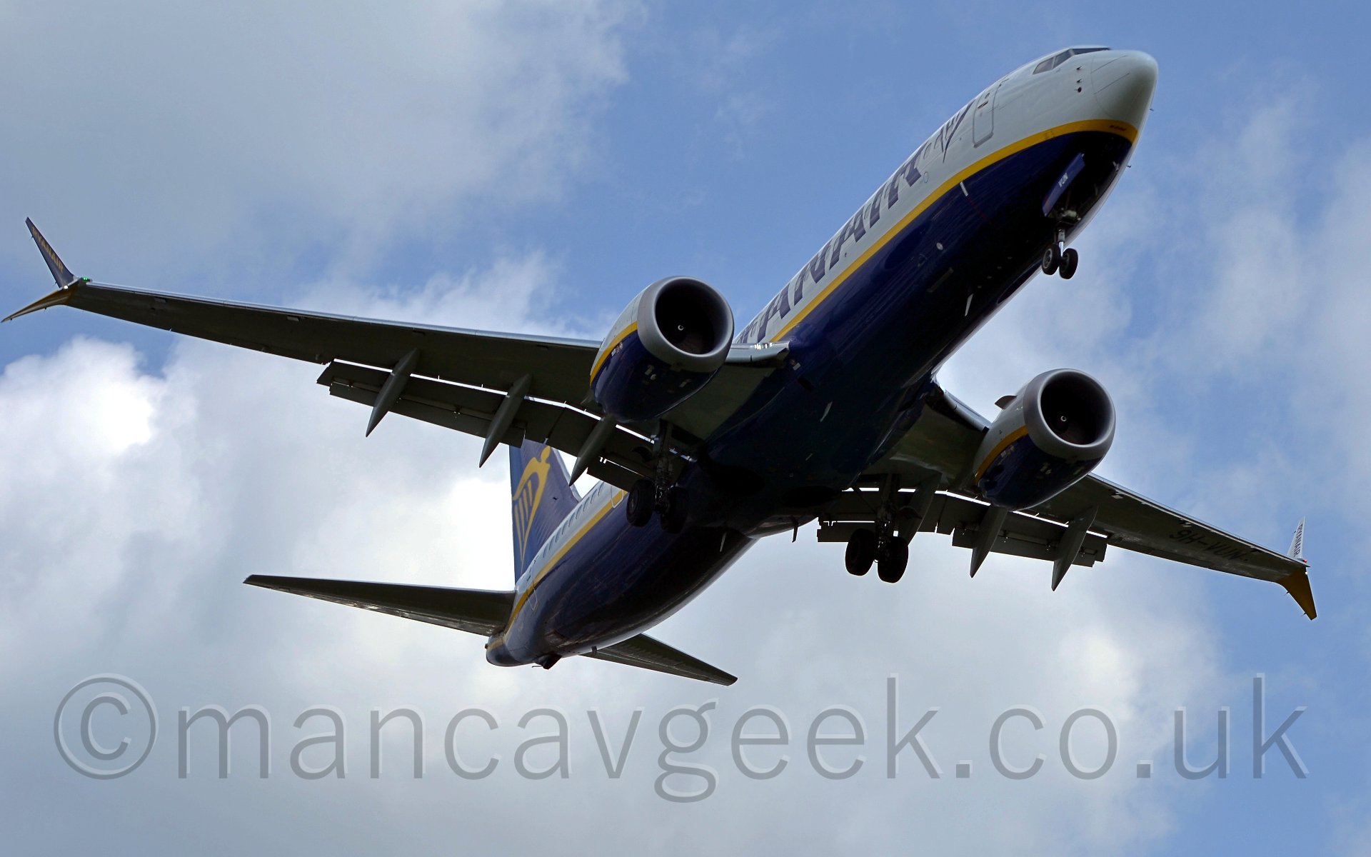 Low side view of a twin engined jet airliner flying from left to right at a very low level, with undercarriage lowered and flaps deployed from behind the wings. The plane has a dark blue belly with white upper section, seperated by a yellow stripe, with the same basic colour scheme on the engines. There are large "Ryanair" titles on the upper forward fuselage. The tail is dark blue, with a yellow winged Irish harp. The wingtips are split, a blue section heading upwards at around 30 degrees, while a yellow portion points about 30 degrees down. Behind the plane, the sky is a mix of piercing blue, and fluffy white.