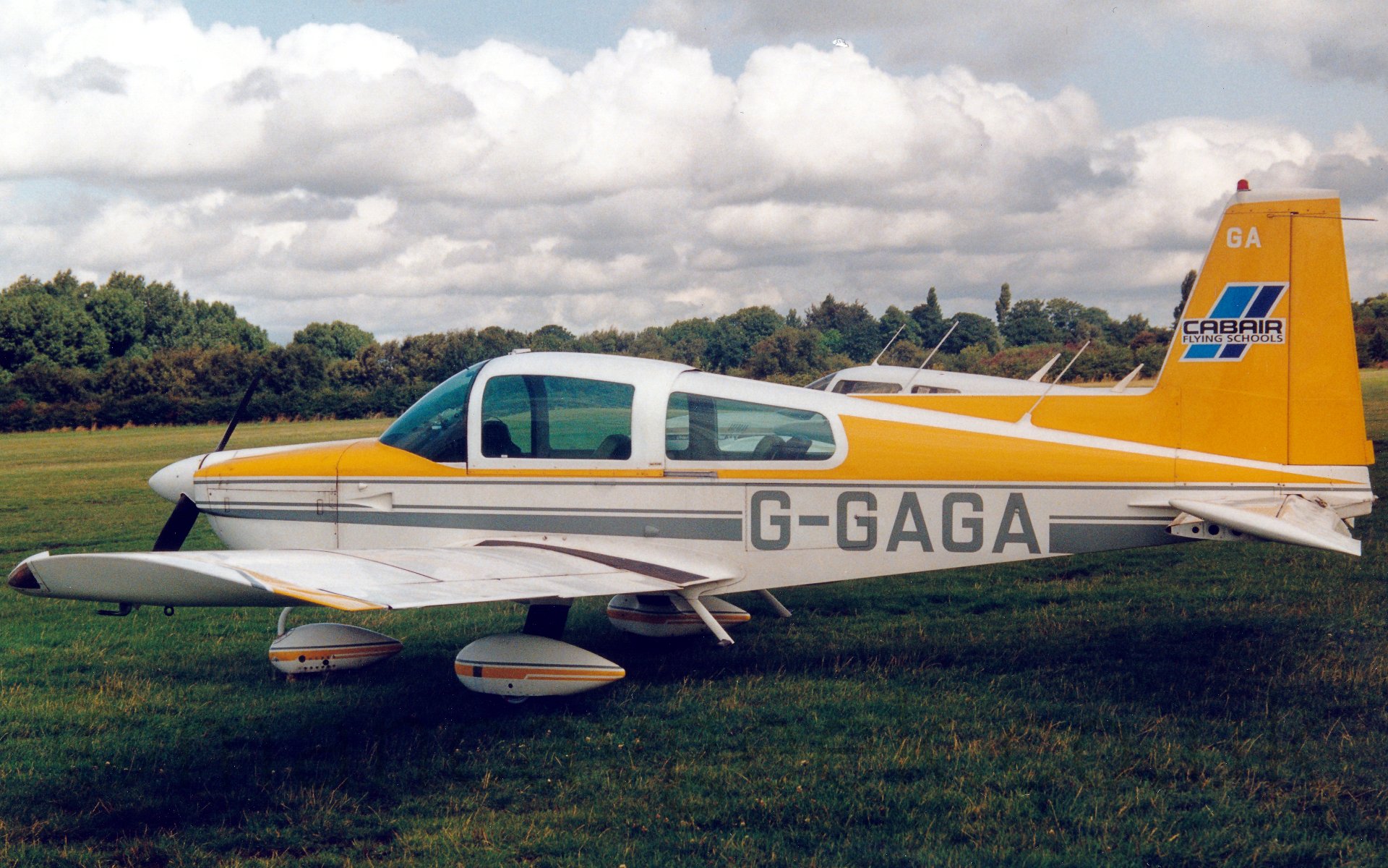 Side view of a single engined light aircraft parked on grass, facing to the left. The plane has a white lower half, with a yellow top, , and a grey stripe in the middle. The registration "G-GAGA" is on the middle of the rear fuselage in grey, while there is a white and blue "Cabair Flying Schools" logo on the tail. There is the top of another light aircraft visible just over the rear fuselage. In the background, grass leads up to a load of bushes at the airfield perimeter, under lots of fluffy white and grey clouds.