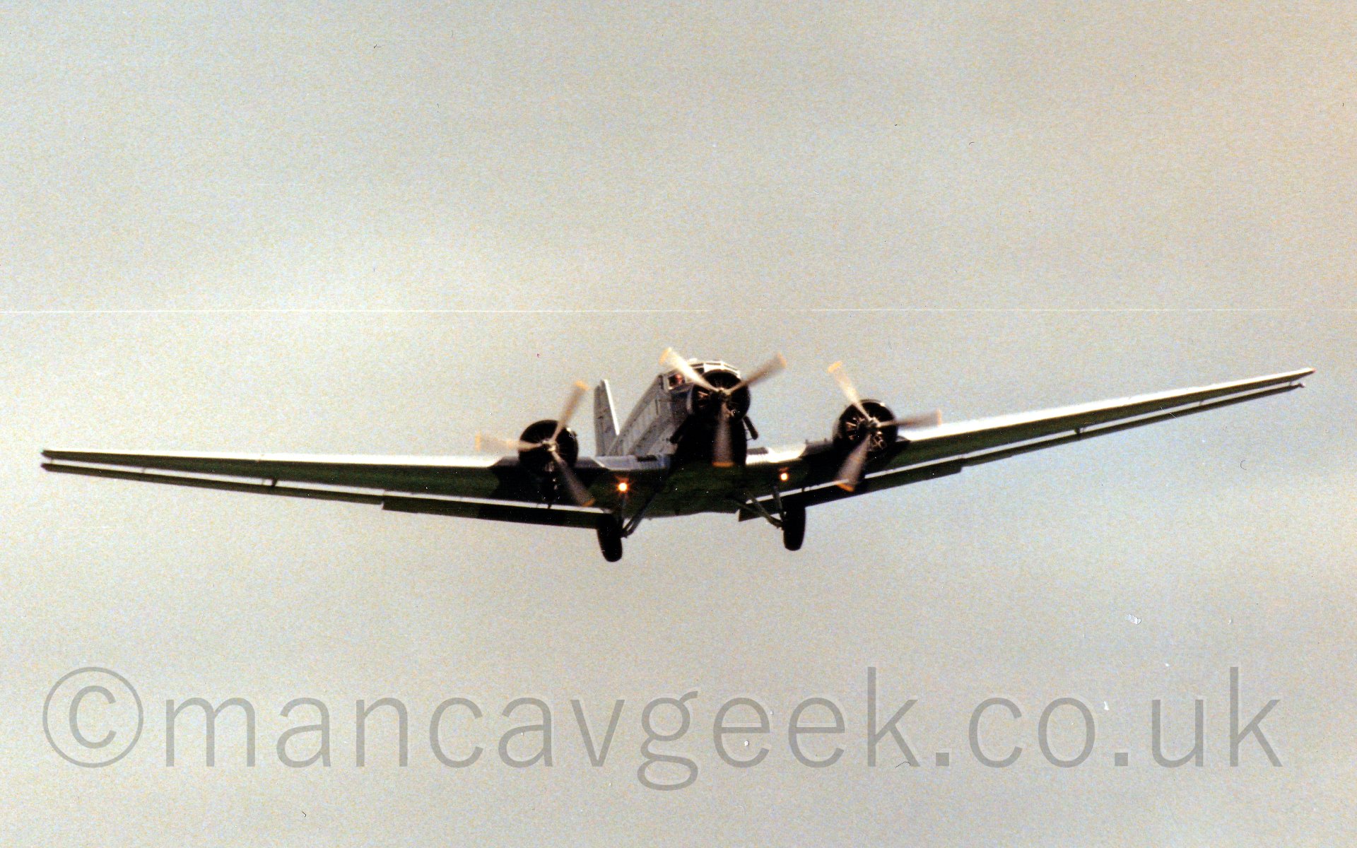 Low front view of a three propellor-engined airliner flying at low level slightly off to the right of the camera.the plane has an engine on each wing and one on the nose, with fixed undercarriage, and with flaps visibly running the full length of the wings. The gun-metal grey plane somehow stands out aginst the grey sky behind it.