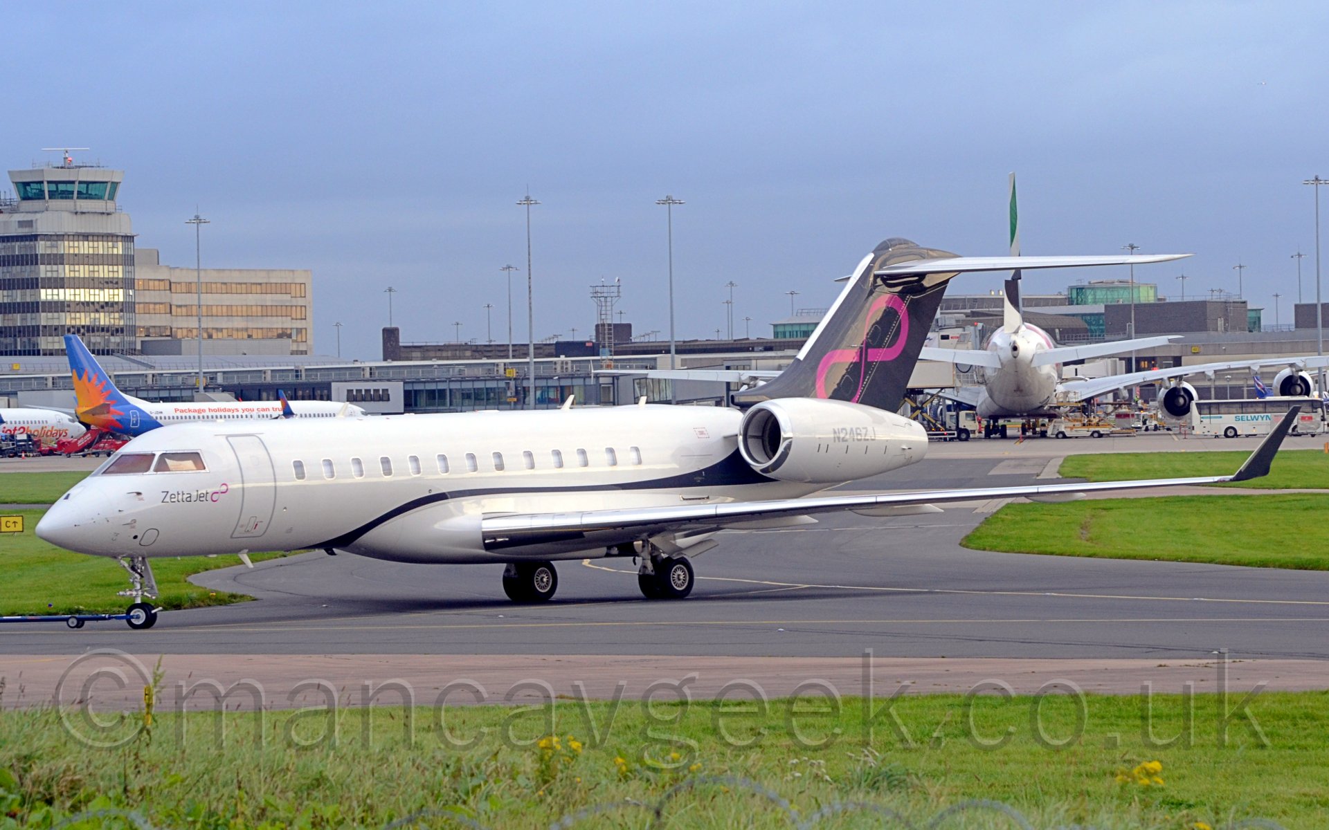 Side view of a large twin engined bizjet facing to the left. The plane is mostly white, with a blue stripe sweeping from below the fuselage just in front of the wing, over the wing, and sweeping upo to cover the entire tail, where there is a large pink infinity symbol. There are small "Zetta Jet" titles along with a small pink infinity symbol on the planes nose, just under the cockpit windows. There is a strip of green grass with wild flowers in the foreground, with more patches of grasson the right and left of the plane in the near background. Further back, there are larger planes parked iun front of a terminal building, with a large building including the control tower on the left of frame. Behind it all, the sky is a pale hazy blue.