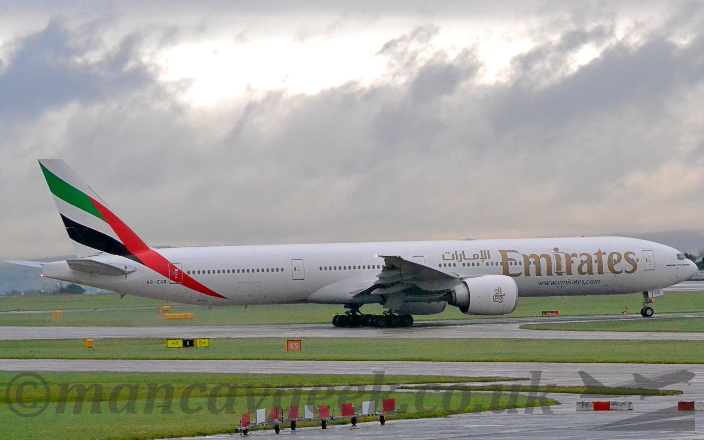 Side view of a twin engined jet airliner moving from left to right. The plane is mostly white, with large golden billboard-style "Emirates" titles in English on the forward fuselage, just ahead of smaller golden tiles in an Arabic script. There is a red line running up from the rear fuselage in to the tail, with green, white, and black stripes coming off it and running across the tail, suggesting the flag of the United Arab Emirates. Large grassed areas crossed by taxiways make up the foreground, with more grass in the background, all under a dramatically grey sky with golden sunlight bursting through at the top.