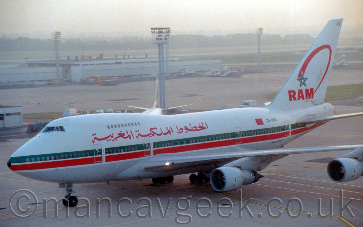 Side view of a very large 4 engined jet airliner taxiing from right to left. The plane is mostly white, with a grey belly, and thick green and red stripes running along the body from the nose, covering the lower deck passenger windows. There is a large, red piece of Arabic script on the upper forward fuselage, transliterating into English as Royal Air Morocco". On the tail, there is the green outline of a 5-pointed star, with a red src for a tail, above the red letters "RAM". In the background is a tall lighting pole looming over the plane, with a sprawling white building and a couple more lighting poles further back, with hazy trees in the background.