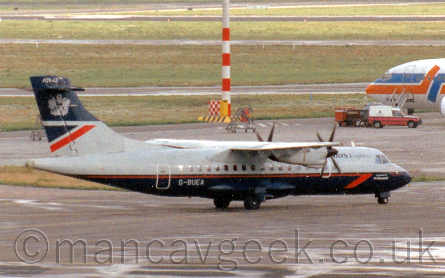 Side view of a T-tailed, high-winged, twin propellor-engined airliner taxiing from left to right. The plane is mostly a dark grey, with a deep blue belly with a thin red stripe running along the body.There are "British Airways Express" titles on the upper forard fuselage, partially obscured by the engines. The tail is grey at the bottom, with a dark blue top, with a dark blue traingle and diagonal red stripe seperating the 2 sectionswith a Royal crest on the top half, and "ATR42" titles right at the top. In the background is a lighting pole with a red and white banded base, and the nose of a larger airliner on the right of the frame, with alternating grass and tarmac areas leading off in to the distance.
