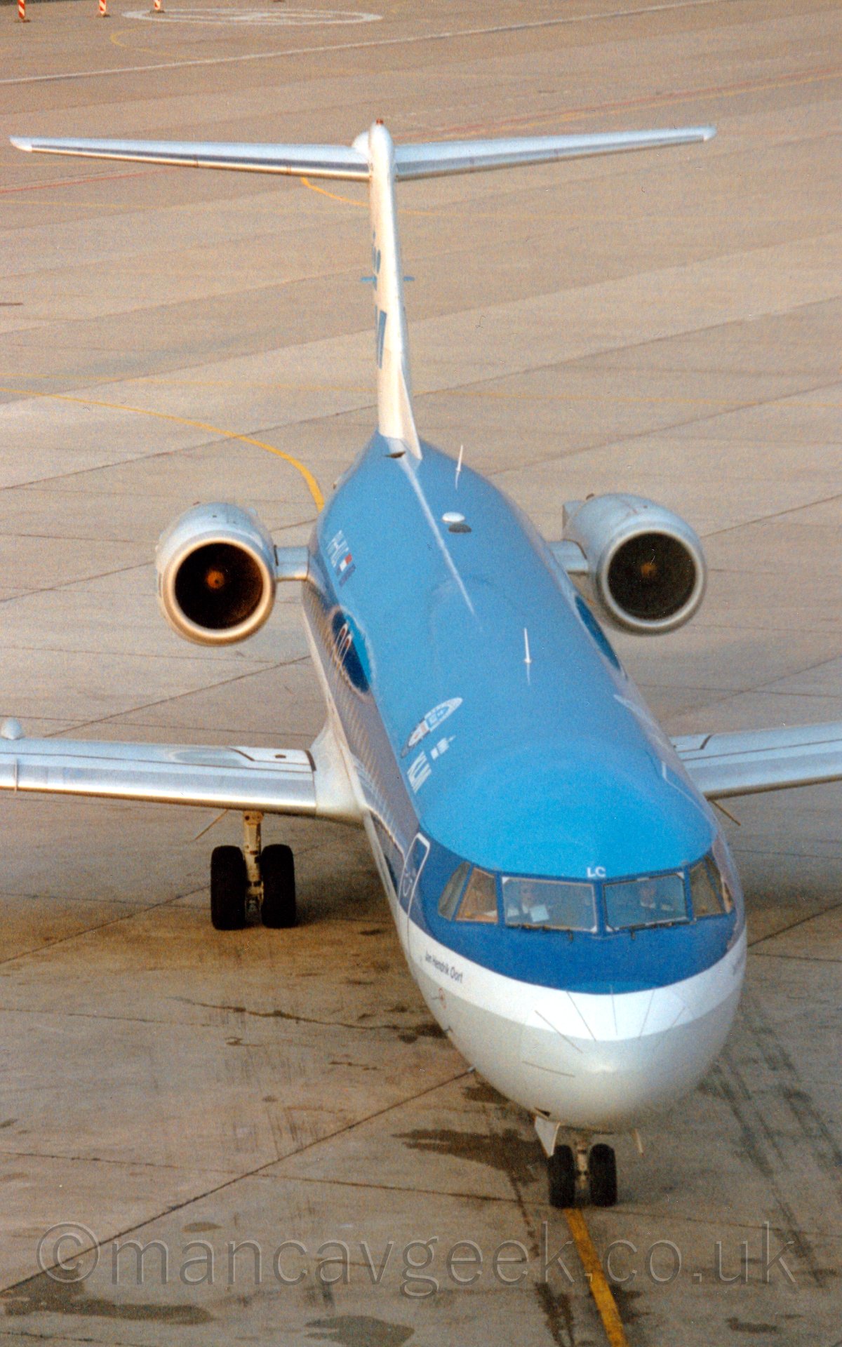 High front view of a t-tailed twin-engined jet airliner taxiing to a spot almost directly below the camera. The plane has a light blue upper section with a grey belly, and thick dark blue and white stripes running the the length of the body with white "KLM" titles under a white crown on the upper forward fuselage.