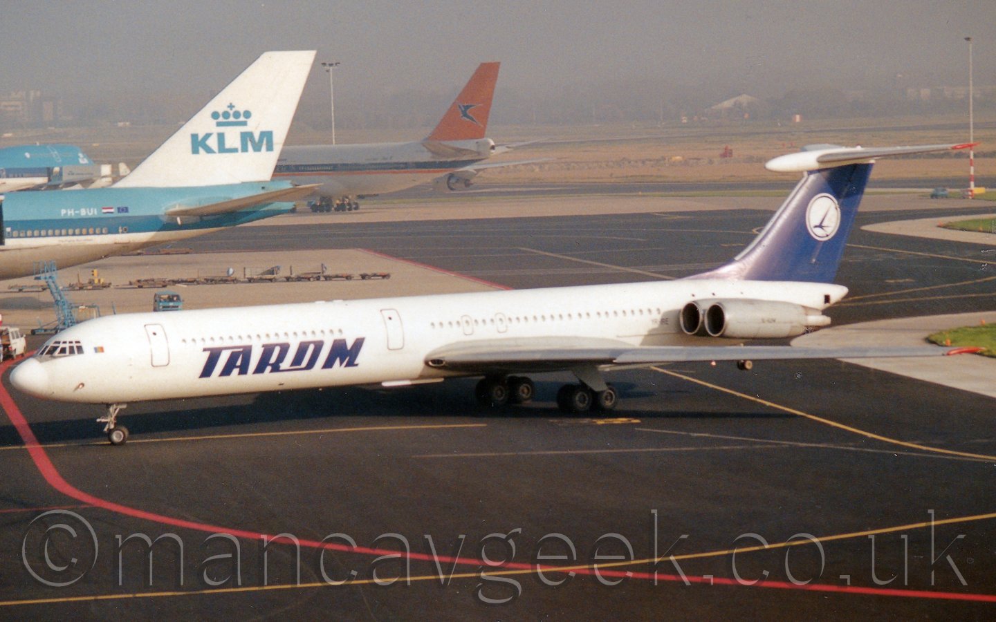 Side view of a T-tailed, 4 engined jet airliner, with the engines mounted on the rear fuselage. The planes body is mostly white, with a black patch over the top of the nose, directly in front of the cockpit, and large "Tarom" titles on the lower forward fuselage. The tail is blue, with a white circle overlaid with the stylised image of a flying bird. In the background, 3 very large 4 engined jet airliners are clustered together in the top left corner, the rest of the image being a view across the airfield before vanishing in to haze in the distance.