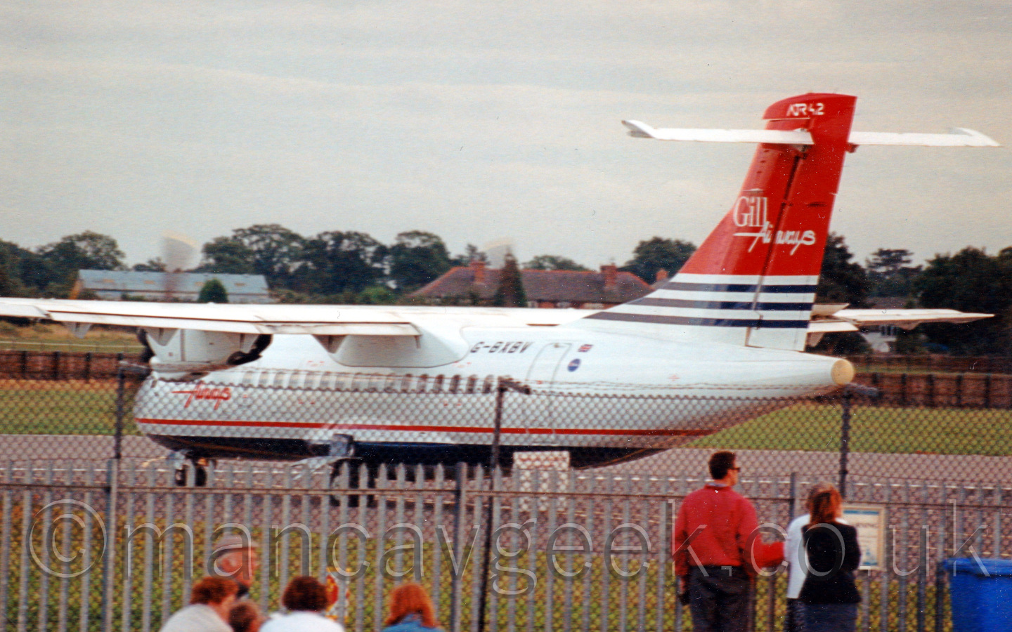 Rear view of a high-winged, T-tailed twin propellor-engined airliner taxiing from right to left and turning away from the camera. The body is mostly white, with a dark blue belly, and a thin red stripe running along the body. The tail is mostly red, with white and blue stripes at the base. There are white "Gill Airways" titles in the middle of the tail, with smaller white "ATR42" titles right at the top. In the foreground, several people are watching through a chain-link fence, while in the background, houses and other buildings are nestled in trees, under a grey sky.