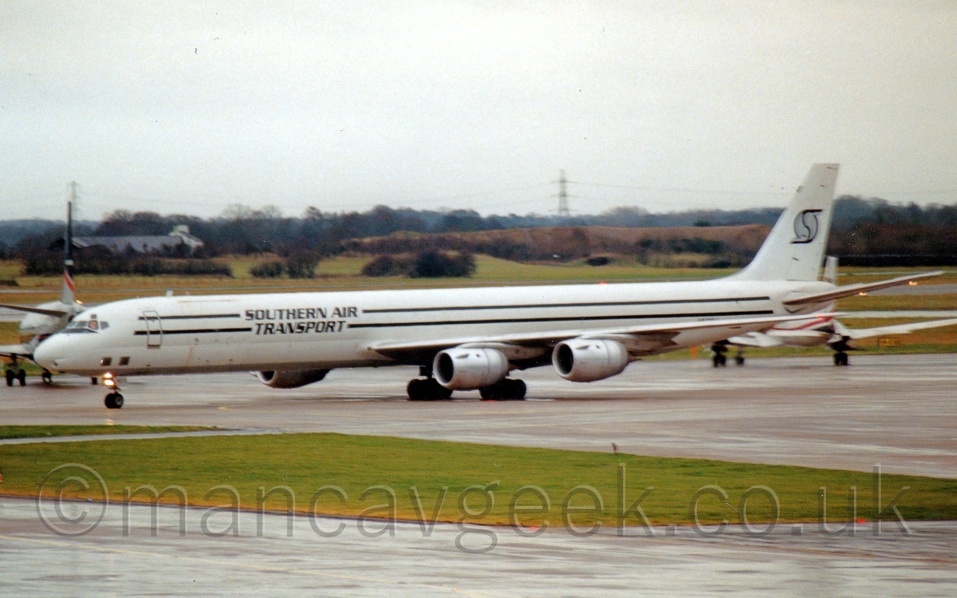 Side view of a rather long and thin 4 engined cargo jetliner taxiing from right to left. The plane is mostly white, with a silver belly, and 2 black stripes running along the top and bottom of the fuselage, with "Southern Air transport" titles overlaid. The tail has a grey circle overlaid with a black letter "S". In the background, a couplke of planes are facing away from the camera, one on each edge of the frame. In the distance, fields and trees lead off i to the distance. under a bright white sky.