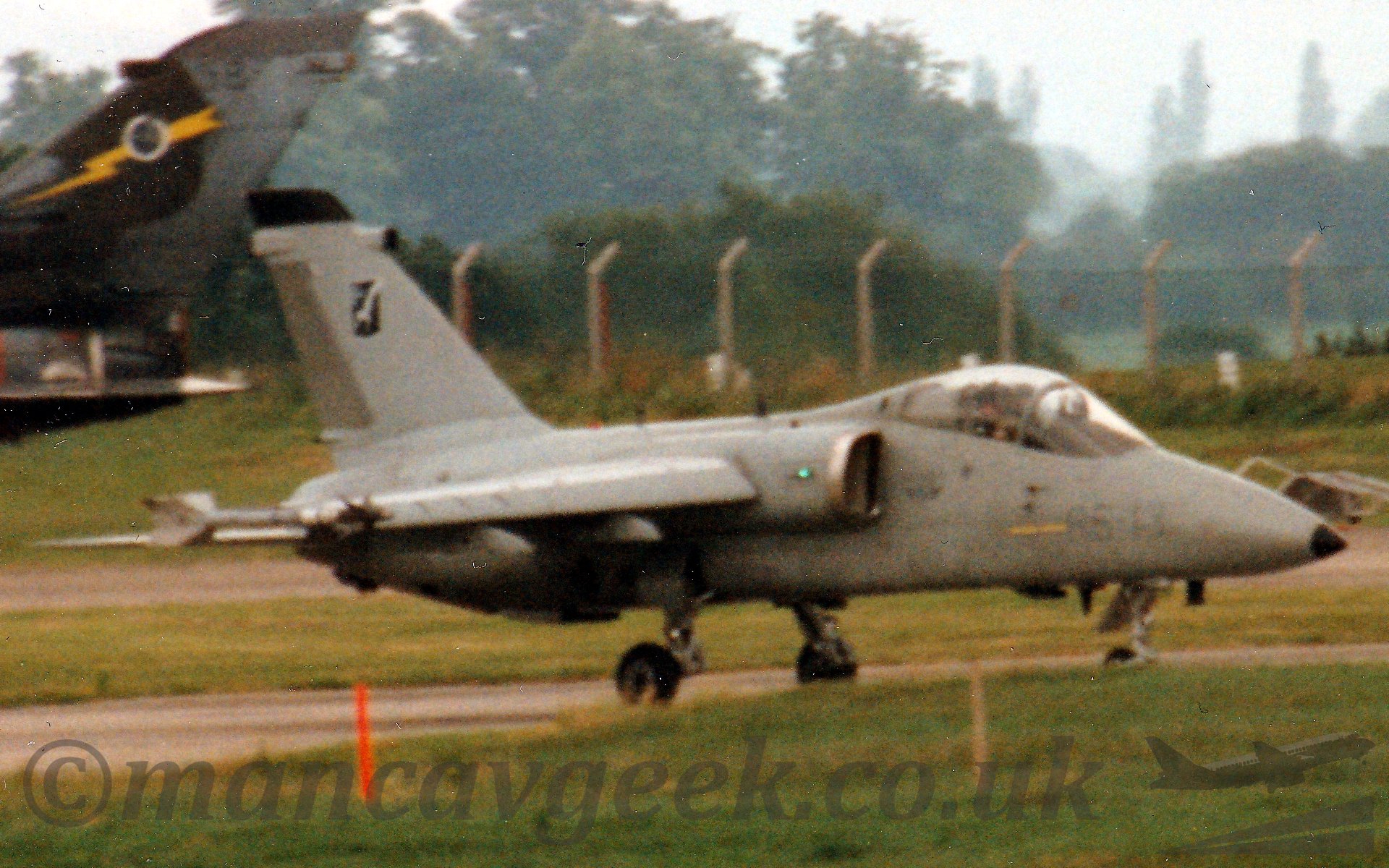 Side view of a single seat, single enginedattack jet aircraft taxiing from left to right. The plane is almost entirely grey, except for the extensively glazed cockpit canopy. There are engine intakes tigh up on the sides of the fuselage, just aft of the cockpit, with the wings coming straight out of the intake. There are missiles carried on the wingtips. The tail has a black badge with a silver eagle flying to the left. In the background, the tail of another plane, with green and grey camouflage and a yellow lightning flash on the tail, is just poking into the frame on the left. There is a chainlink fence mounted on conctrete posts running across most of the frame, with trees and bushes behind it, all under a grey sky. The photo is, unfortunately, rather grainy, so a lot of detail has been lost.
