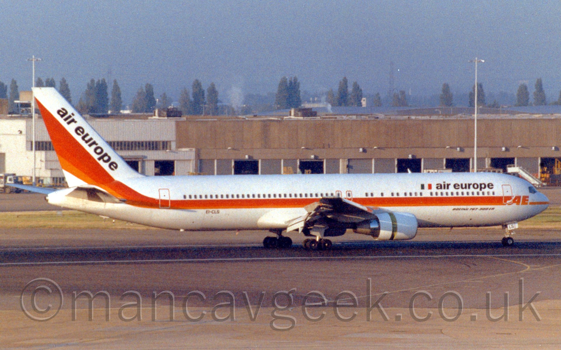 Side view of a twin engined jet airliner travelling from left to right, with flaps fully extended behind the wing, and the thrust reversers open on the engines, suggesting it has just landed. The plane is mostly white, with a red and orange stripe running along the body from the nose and sweeping up into the tail, with the letters "AE" overlaid at the front, between the cockpit and the forward door. There is an italian flag (Green, white, and red vertical bars) and "Air Europe" titles on the forward upper fuselage, just aft of the forward door. There are also diagonal "Air Europe" titles on the tail. Behind, there is a large brown cargo hangar, with trees and buildings vanishing in to haze in the distance, under a leaden grey sky.