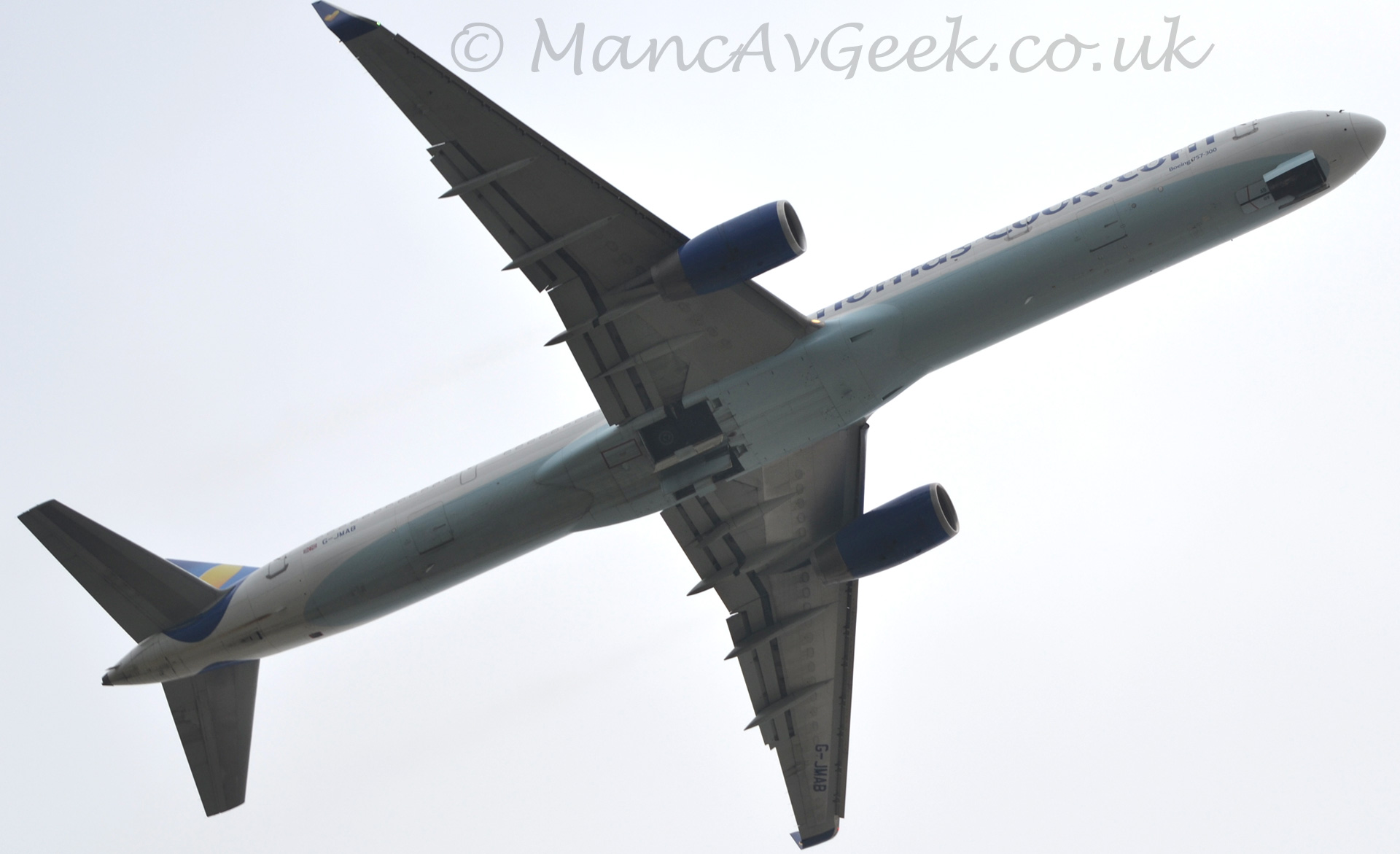 View from almost directly below of a twin engined jet airliner flying from left to right at low level, with the landing gear being stowed, and flaps deployed from the rear of the wings. The plane is mostly white, with a duck-egg blue belly, and large dark blue "Thomas Cook.com" on the forward fuselage. The tail is blue, with a yellow splodge. The sky behind it is a flat grey.