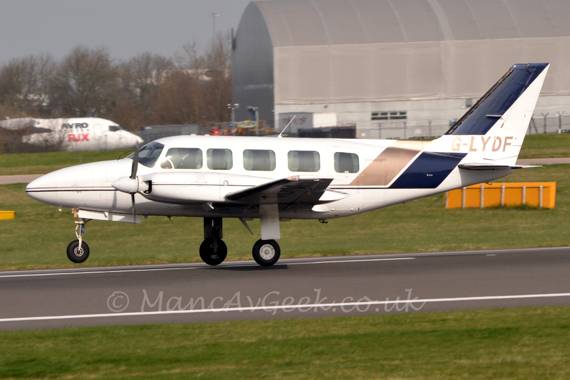 Side view of a twin propellor engined business aircraft moving from right to left, with the nose lifted slightly off the ground as it rotates off the runway. The plane is mostly white, with a thin blue and gold stripe running along the body from the nose, widening behind the cabin windows, before sweeping up to occupy most of the tail. The registration "G-LYDF" is on the tail in large golden letters. In the background is a large grey-brown hanger, behind which is a white jet airliner with "Avro RJX" titles on the forward fuselage.