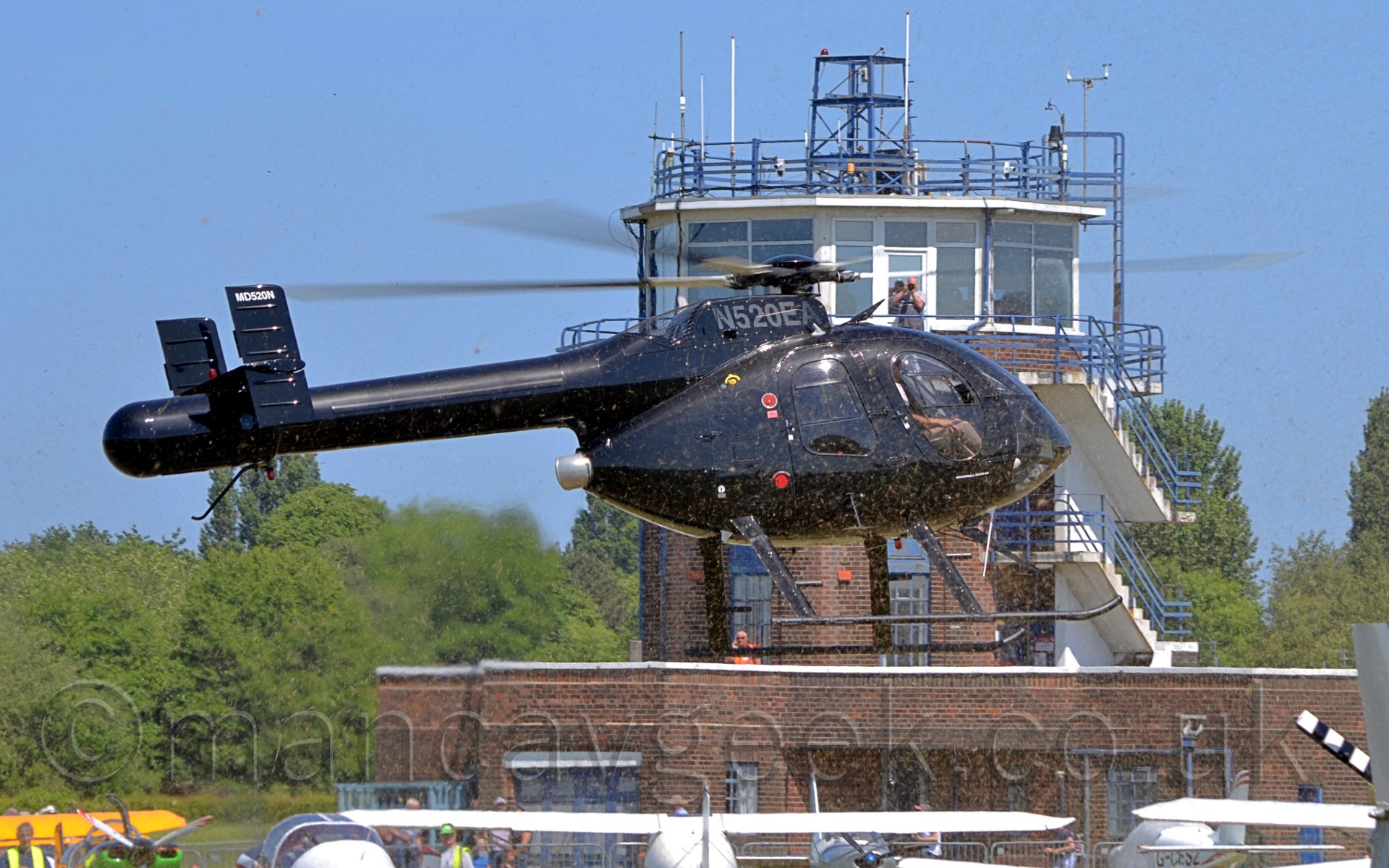 Side view of a helicopter hovering several feet above the ground, facing to the right. The helicopter is completely black, with the registration "N520EA" on the engine on top of the fuselage. In the background is a brown building with a fully glazed room occupying the whole top floor, with several antennae on top. A man is standing on a blacony taking a photo of this helicopter, but looks as if he is taking a photo of me and my camera.