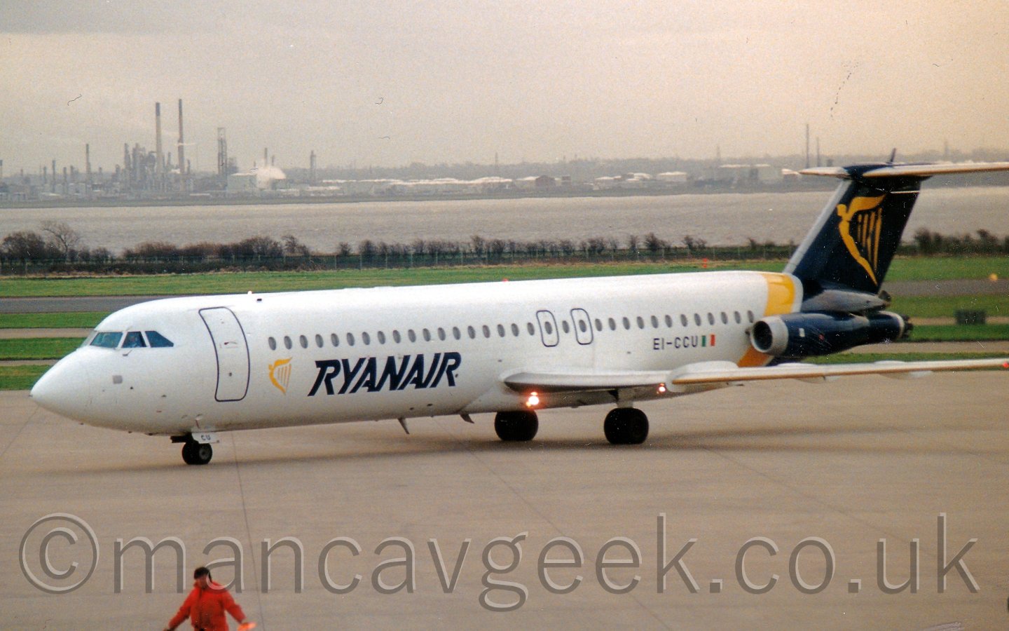 Side view of a T-tailed twin engined jet airliner with it's engines mounted on the rear fuselage. The plane is mostly white, with a diagonal yellow band around the rear fuselage, the end of the fuselage and tail being filled with a dark blue, with a winged golden Irish harp. There is another, smaller, harp on the lower forward fuselage, withlarge dark blue "Ryanair" titles. In the background, large areas of grass lead up to a wide river, with large scale industrial facilities on the far bank.