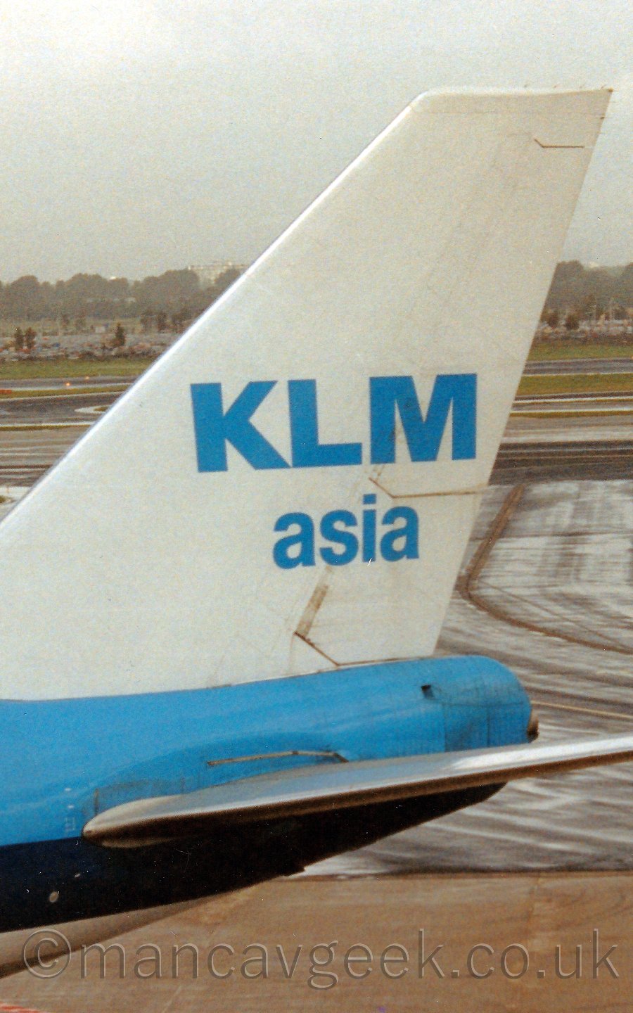 Close up of the tail of a very large 4 engined jet airliner, facing to the left. The plane is mostly blue, with a white tail, which has blue "KLM Asia" titles. behind is a view across the airfield, to trees and buildings vanishing in to haze.