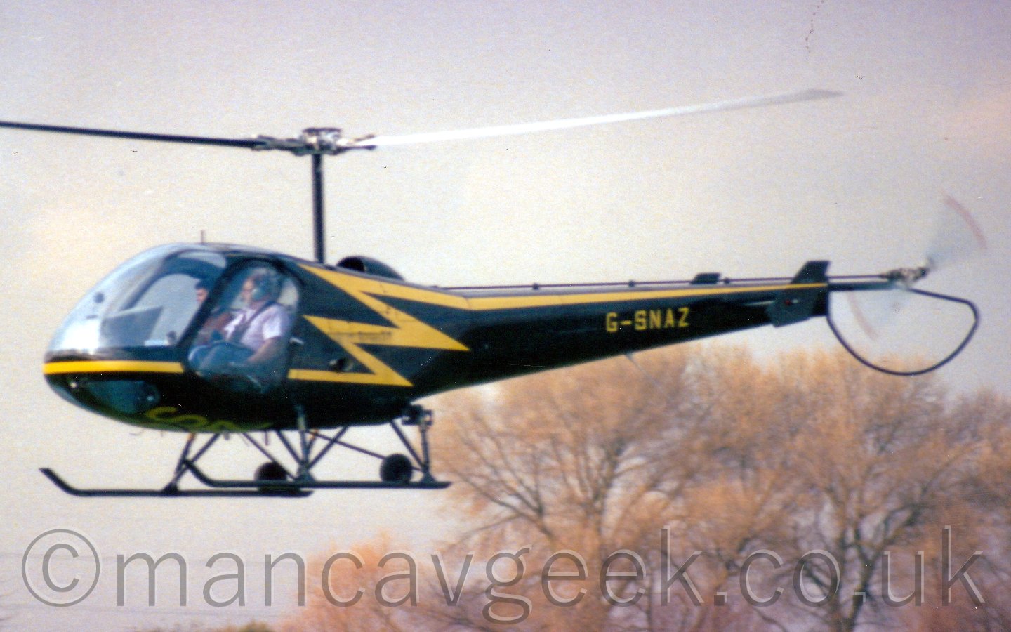 Side view of a helicopter flying from right to left at low altitude. the helicopter is black, with a yellow stripe running along the body, turning into a lightning boltas it moves to the top, then running straight along the tail boom to the tail rotor. The highly glazed cockpit has two people inside. Behind, there are some trees behind the helicopter, with grey sky beyond that.