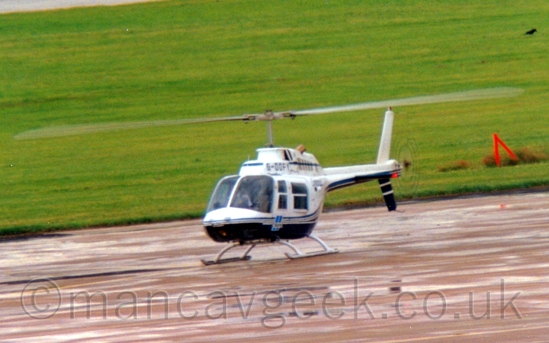 Side view of a helicopter on the ground with both it's main and tail rotors turning. The body is mostly white, with dark blue lower surfaces, the 2 sections seperated by thin light blue and brown stripes running all the way from the nose to the tail. In the background, the apron the helicopter is parked on gives way to a large expanse of green grass which fills the frame.