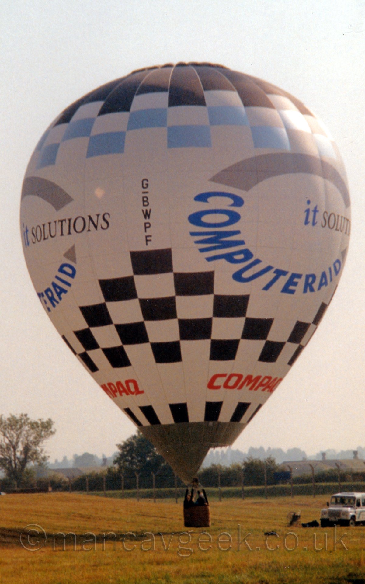 Side view of a hot air balloon taking off from a grassed area, There are 2 people in the wicker basket at the bottom, just a few feet above the ground. The balloons canopy is mostly white, with a black and white checkerboard pattern at both the top and bottom, with additional blue checkerboard panels at the top. There are red "Compaq" titles towards the bottom, and blue "Computeraid" titles forming the lower half of a circle in the centre, the rest of the circle being a grey line., with blue and grey "it solutions" titles in the middle. There is a whiteoff-road vehicle parked off to the right of the frame. In the background, a chainlink fenceruns across the middle of the frame, with trees and bushes behind it, with grey sky completeing the image.