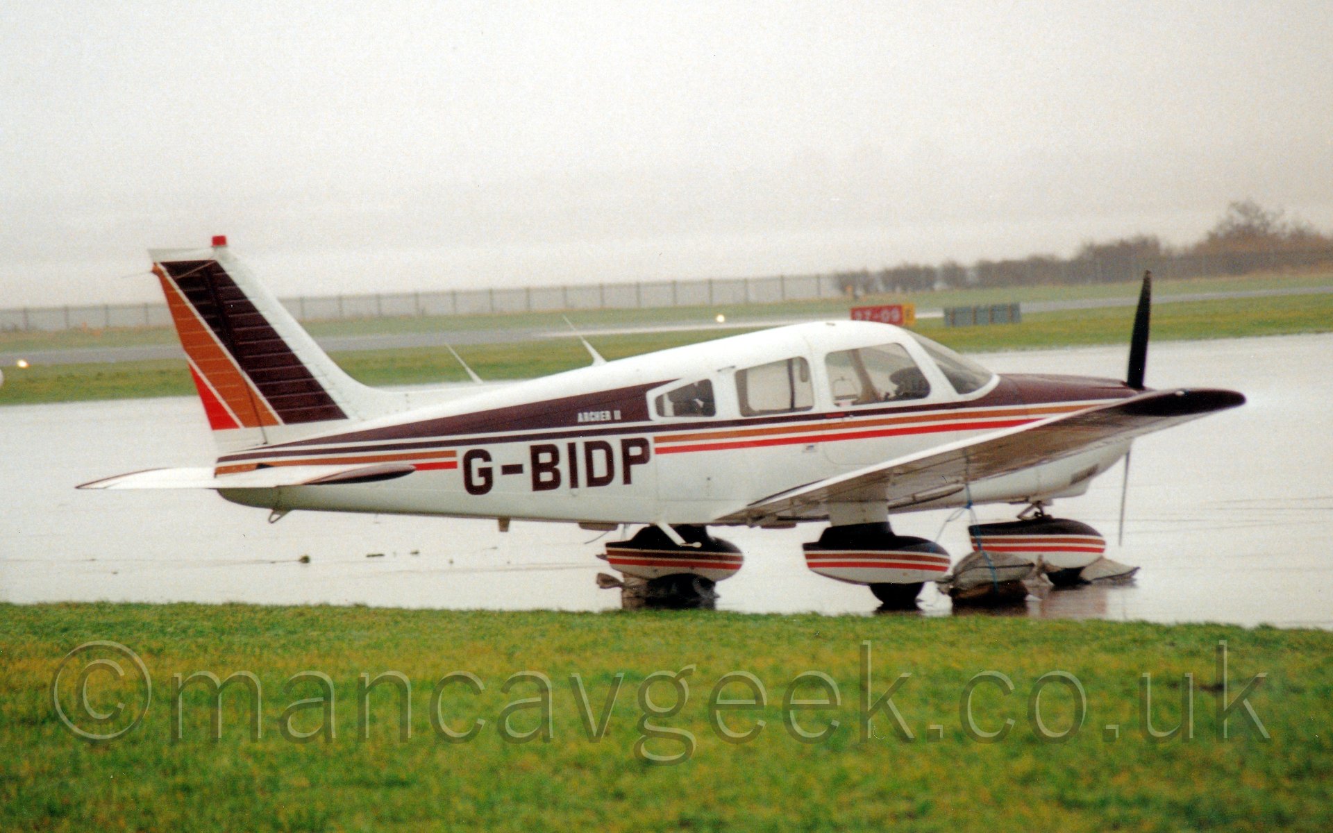 Side view of a single engined light aircraft parked facing to the right. The plane is mostly white, with a thick brown stripe covering most of the upper fuselage and thin orange and red stripes running along the middle of the body. The same design covers the spats over the fixed undercarriage, as well as on the tail. In the background, the rain-covered apron stretches off into the distance, giving way to grass which runs off to the airfield perimeter, vanishing in to the rain.