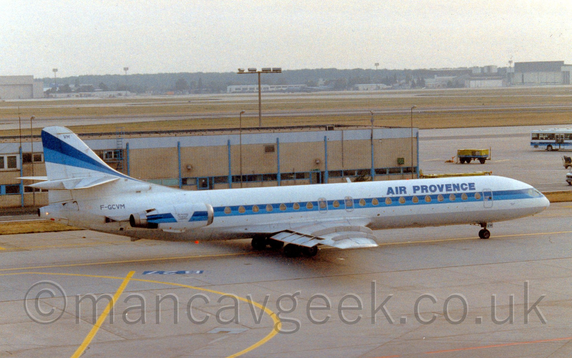 Side view of a twin engined jet airliner with the engines mounted on the rear fuselage taxiing from left to right. The plane is mostly white, with a thick light and dark blue running along the fuselage, covering the passenger cabin windows, and over the engines, with another diagonal stripe running up the tail. There are dark blue "Air Provence" titles on the upper forward fuselage. In the background, there is a low, sandy brownbuilding that extends 2/3rds of the way into the frame from the left, with a single deck bus on the right, wearing a similar colour scheme to the plane. In the far distance, more buildings are visible on the far side of the airport, with rows of trees behind that, fading into haze.