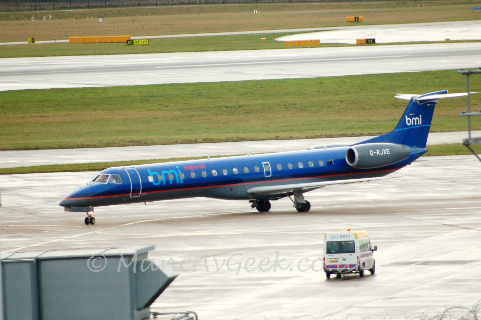 Side view of a twin engined jet airliner taxiing from right to left. The plane is mostly dark blue, with a dark grey belly, seperated by a thin red stripe. There are large, light blue "bmi" titles, followed by smaller red "regional" titles on the upper forward fuselage. The engines are grey, with the registration G-RJXE in white, attached to the side of the rear fuselage. The tail is the same dark blue, with a light blue stripe below white "bmi" titles. In the foreground there is a large grey ventilation ducting on the lower left of the frame, with a white van with "security" titles on the upper part of the rear doors. In the background, alternating stretches of grass and tarmac stretch off into the distance.