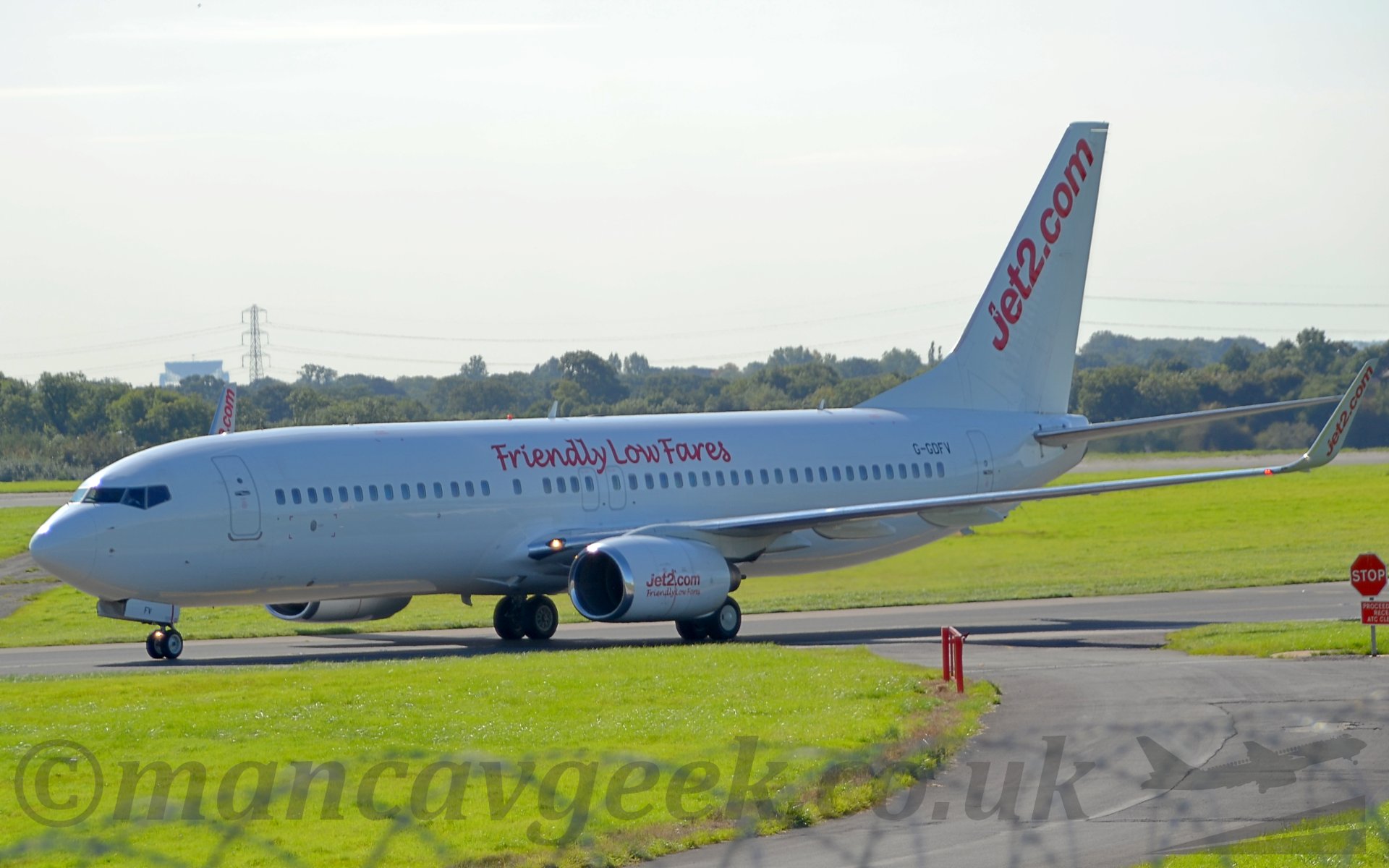 Side view of an all white twin engined jet airliner taxiing from right to left. There are red "Friendly Low Fares" titles in the upper fuselage over the wings, diagonal red "Jet2.com" titles on the tail and upturned wingtips, and both sets of titles on the engine pods. In the background, trees and bushes mark the airfield perimeter, running off into the distance. On the horizon, the dish of a radio telescope can be seen over the planes cockpit on the right of the frame. Above, the sky is a hazy bright white.