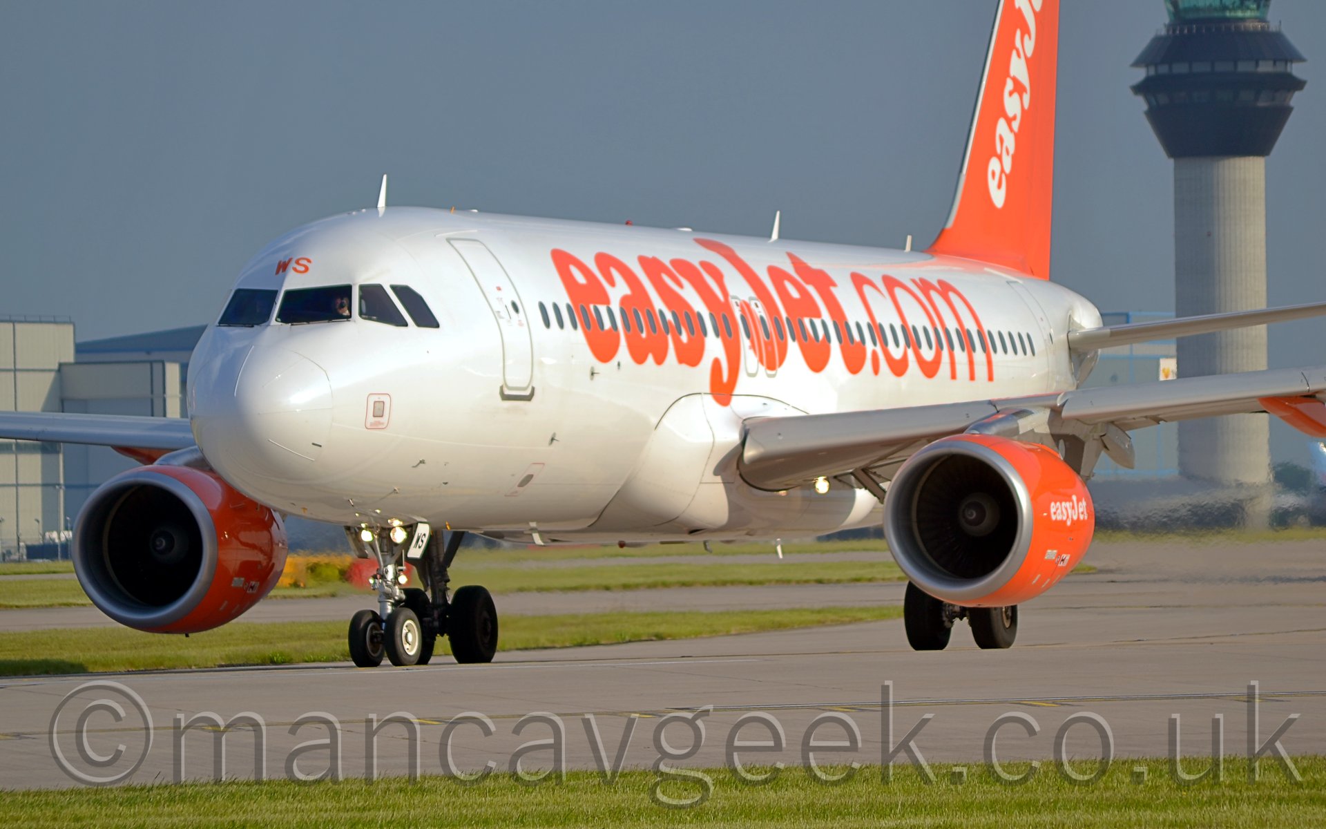 Side view of a twin engined jet airliner taxiing to the left of the camera, turning further to the left. The plane is mostly white, with large billboard-style orange "easyJet.com" titles on the fuselage, The tail is a bright orange, with diagonal white "easyJet" titles, with the engines also being orange with white titles. In the background, a large grey hangar is visible over the right wing, whileanother large grey hangar and a tall control tower is visible over the right wing. The sky is a hazy blue-grey.