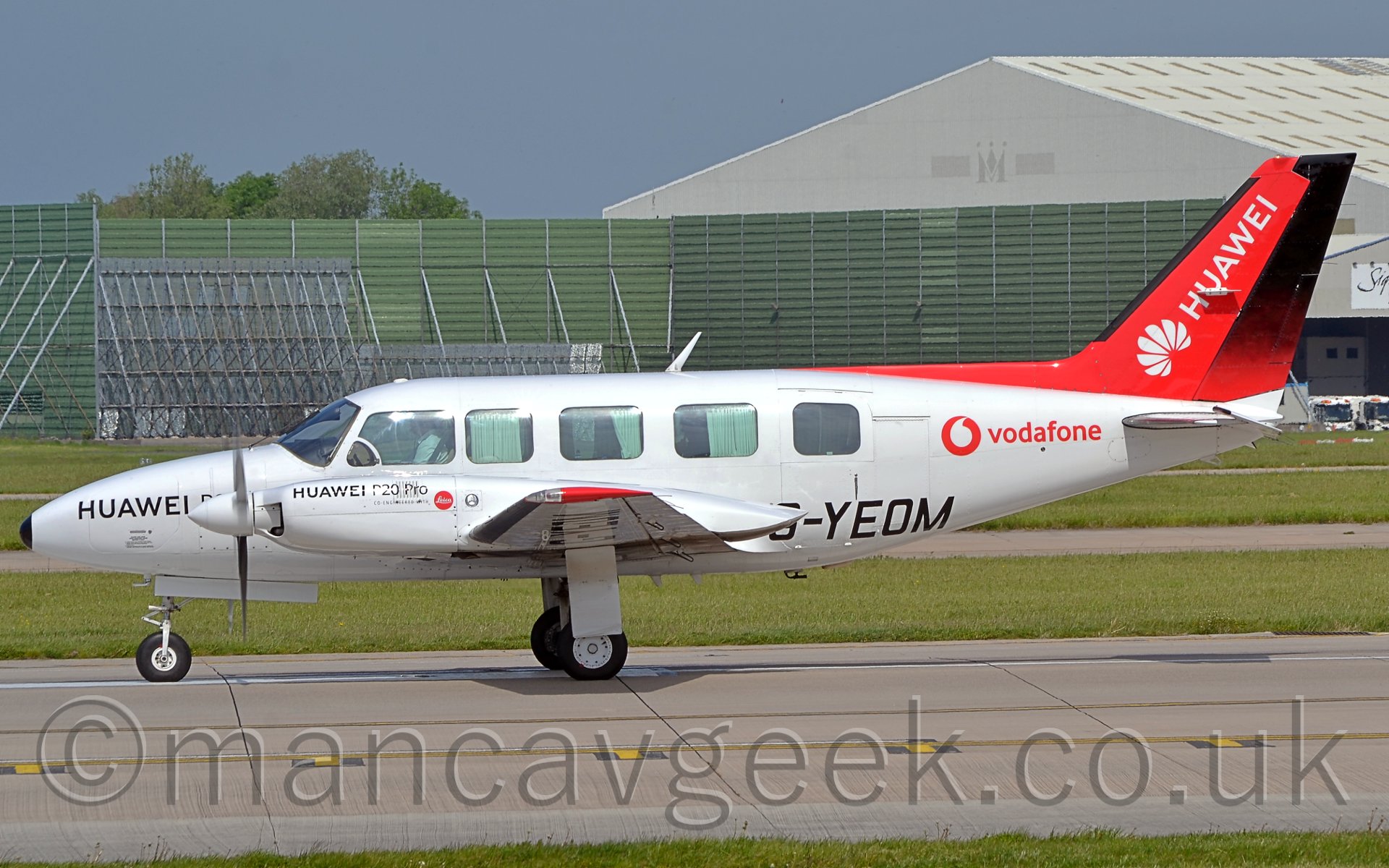 Side view of a twin propellor-engined light business aircraft parked facing to the left. The plane is mostly white, with black "Huawei P20 Pro" titles on the nose and engine cowlings, the registration "G-YEOM" in black on the lower rear fuselage and under the left wing, and a red circle with a white apostrophe in the center, next to red "Vodafone" titles on the upper rear fuselage. The tail is red, with diagonal white "Huawei" titles. In the background, a large, green, open square metal frame can be seen, in front of a large grey hangar on the right of the frame. Above, the sky is a light blue-grey.