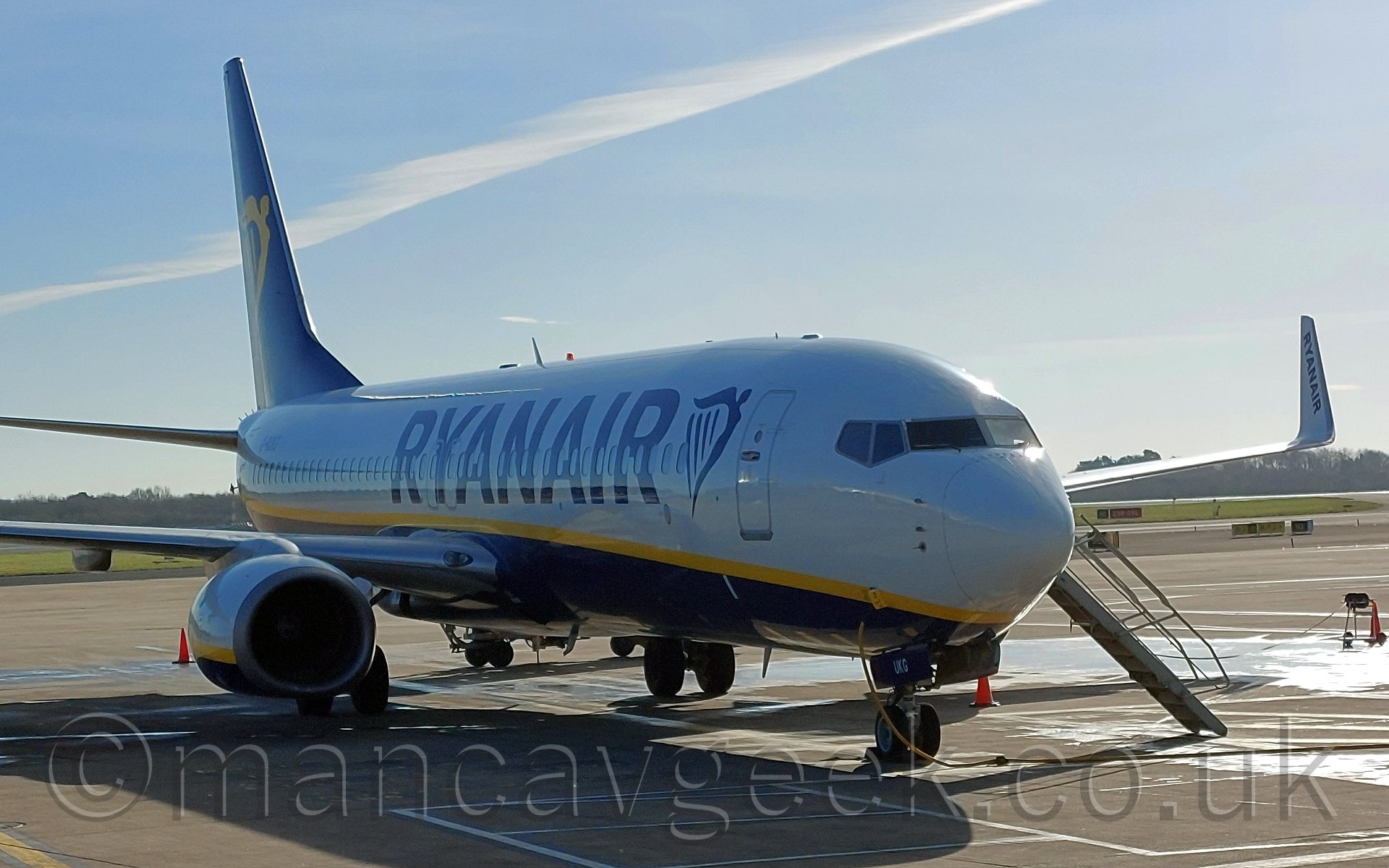 Slight side view of a twin engined jet airliner parked almost nose on to the camera, pointing slightly off to the right. The plane is mostly white, with a dark blue belly, with a thin yellow line seperating the 2 sections. There are large, dark blue "Ryanair" titles on the forward fuselage, next to a dark blue winged Irish harp, the same harp being on the dark blue tail in yellow. There is a built in set of stairs coming from the forward door on the other side of the plane., and a thick yellow cable plugged into a hatch in the planes nose, just forward of the nosewheel. Behind, trees mark the airfield perimeter, giving way to a bright blue sky with a single line of cloud. bright sunlight is coming from off to the right of the frame, casting strong shadows on the ground and leaving some of the planes edges sparkling. Not bad for a photo taken with a cellphone.