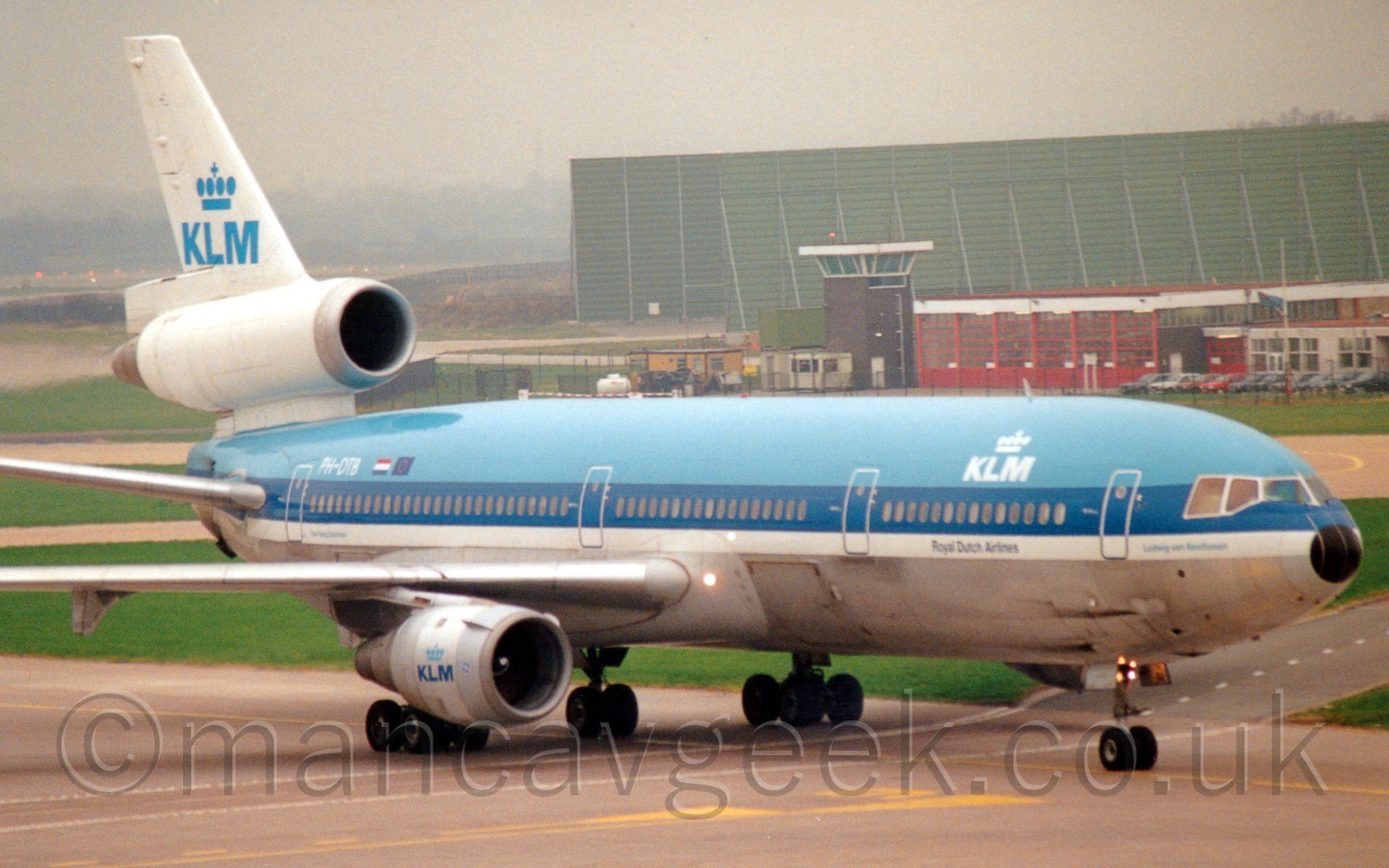 Side view of a 3 engined jet airliner taxiing from left to right. The plane is light blue on the top, with a silver belly, , and a thick darker blue stripe and thinner white stripe in the middle. There are white "KLM" titles under a stylised white crown on the forwar fuselage, with a similar, larger blue crown over blue "KLM" titles on the tail, as ell as on the white engine pods. Behind, there is a low brown building with red-framed doors and windows, with a short brown tower with glazed section on the roof, behind which is latrge green metal structure. Above it all, the sky is a depressingly dreary grey.