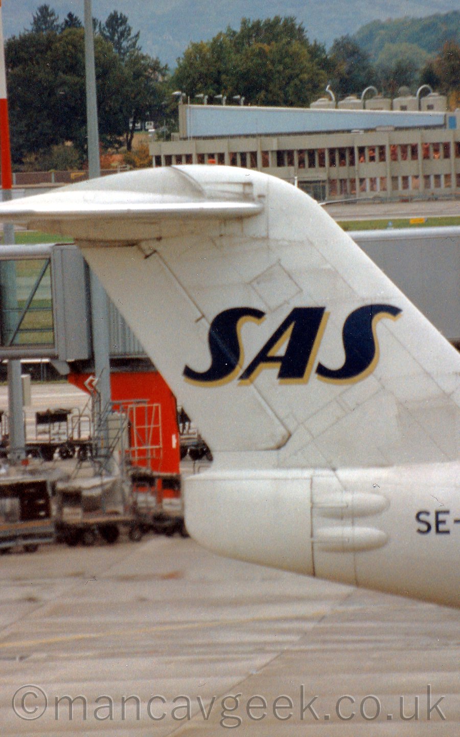 Close up of the tail of a twin engined jet airliner taxiing from left to right. The body and tail are mostly white, with dark blue "SAS" titles with a gold drop shadow on the tail. Behind, a grey airbridge is visible, not attached to anything, with a grey building with and angled blue roof in the far distance, in fron t of some trees.