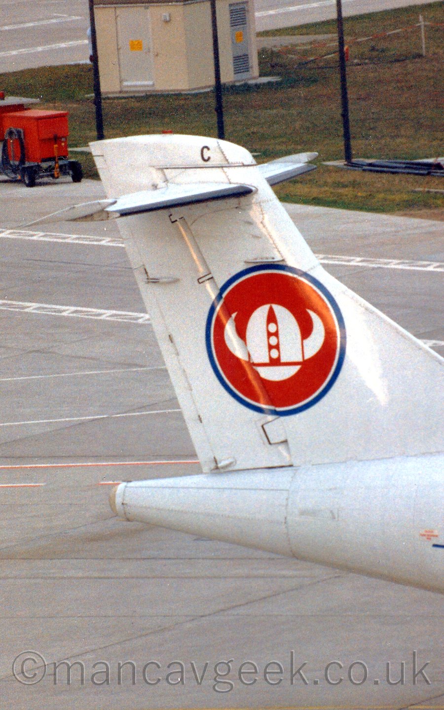 Closeup of the tail of a T-tailed propellor-engined airliner parked facing to the right. The plane is mostly white, with a red circle outlined in blue and overlaid with a white, horned viking helmet.