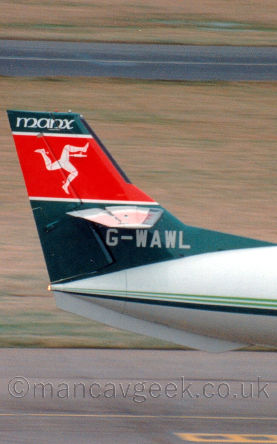 Close up of the tail of a twin propellor-engined airliner taxiing from left to right. the plane is mostly white, with a dark and light green stripe running along the body. The tail is a dark green, with a thick red stripe in the middle carrying 3 bent armoured legs all connected at the hip and Running in a circle The word "Manx" is painted on the top of the tail in white, with the registration "G-WAWL" at the base. In the background, yellowing grass leads off into the distance.