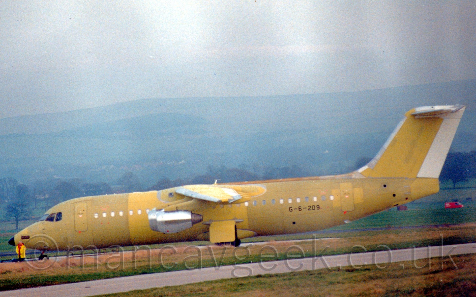 Side view of a high-winged, 4 engined jet airliner being towed from left to right. The plane is covered in a yellowish-green paint, with only the rear of the tail painted white, and silver-coloured engine pods hanging below the wings. There is a person in a yellow hi-viz jacket standing by the plane's nose cone. In the background, trees line the airfield perimeter, with a hill rising in the distance, slowly vanishing in to haze.