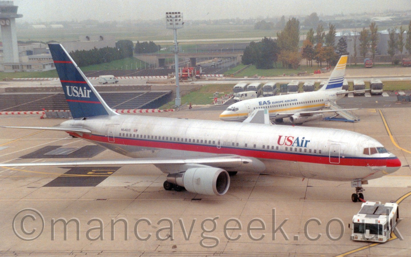 Side view of a twin engined jet airliner being pushed back from it's stand, facing to the right. The plane is mostly in a bare metal finish, with a thick red stripe running along the body, alongside a very narrow dark blue stripe. There are "USAir" titles on the upper forward fuselage in blue and red. The tail is dark blue, with thin red pinstripes, running from front to back, and white "USAir" titles. There is a red pole attached to the nose wheels, the other end of which is attached to a white tug aircraft, which is pushing it backwards. The nose cone is obviously originally from another plane, the colours being very slightly mis-matched and mis-aligned. In the background, a smaller, white twin engined jet airliner is parked facing to the left, in front of a row of buses, next to a tall lighting pole. Trees fill the middle-distance, with large grassed areas and more trees vanishing in to haze in the far distance, under a hazy grey sky.