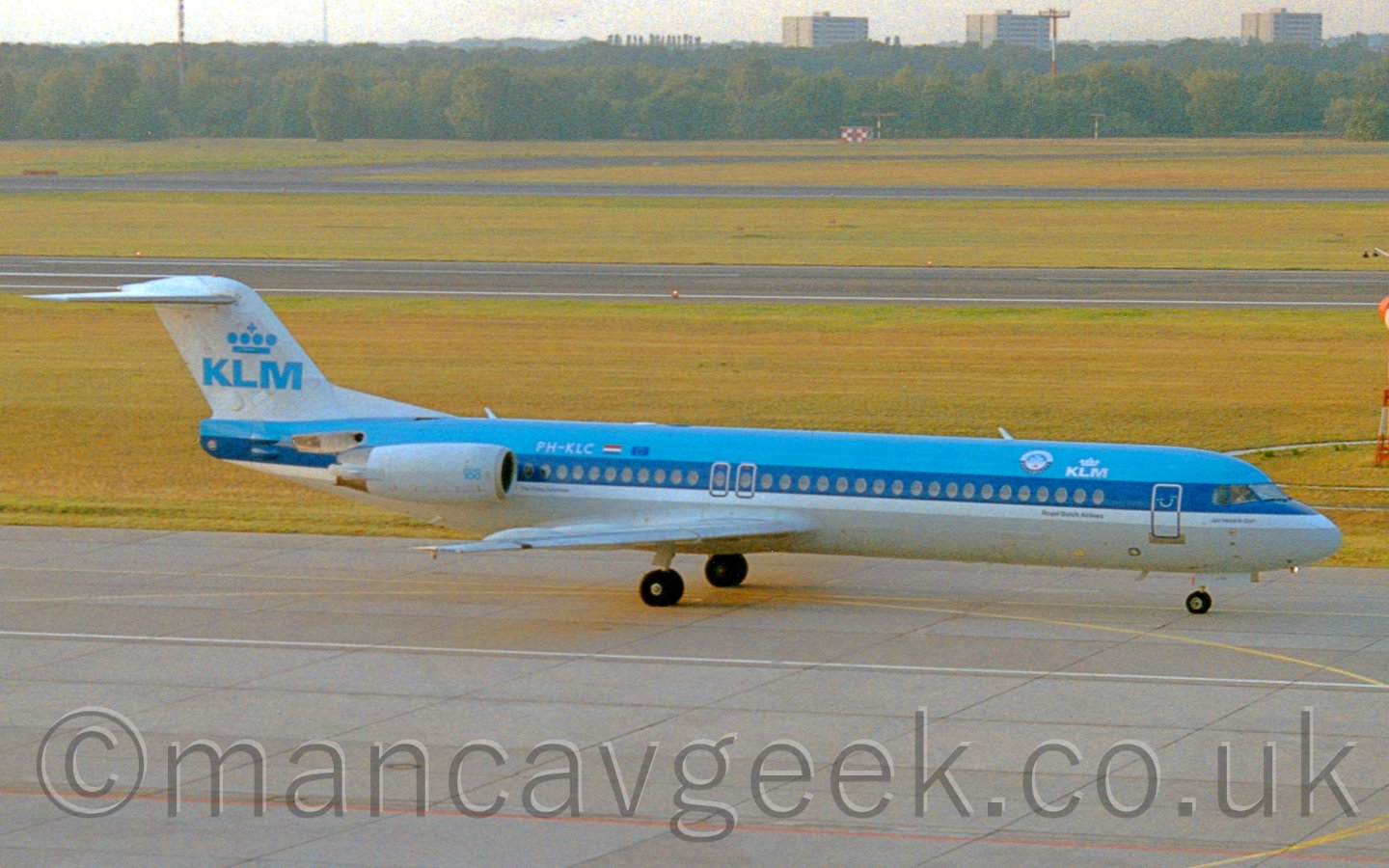 Side view of a twin engined jet airliner with the engines mounted on the rear fuselage taxiing from left to right. The plane has a grey belly and light blue top, with a thick dark blue and thin white stripe seperating the 2 sections. There are small "KLM" titles under a stylised white crown on the upper forward fuselage, with a larger blue "KLM" titles under a stylised blue crown on the tail. There are blue "Fokker 100" titles on the front of the engine pods. In the background, large grass areas are divided by black taxiways and runways, leading off to ranks of trees at the airfield perimeter.