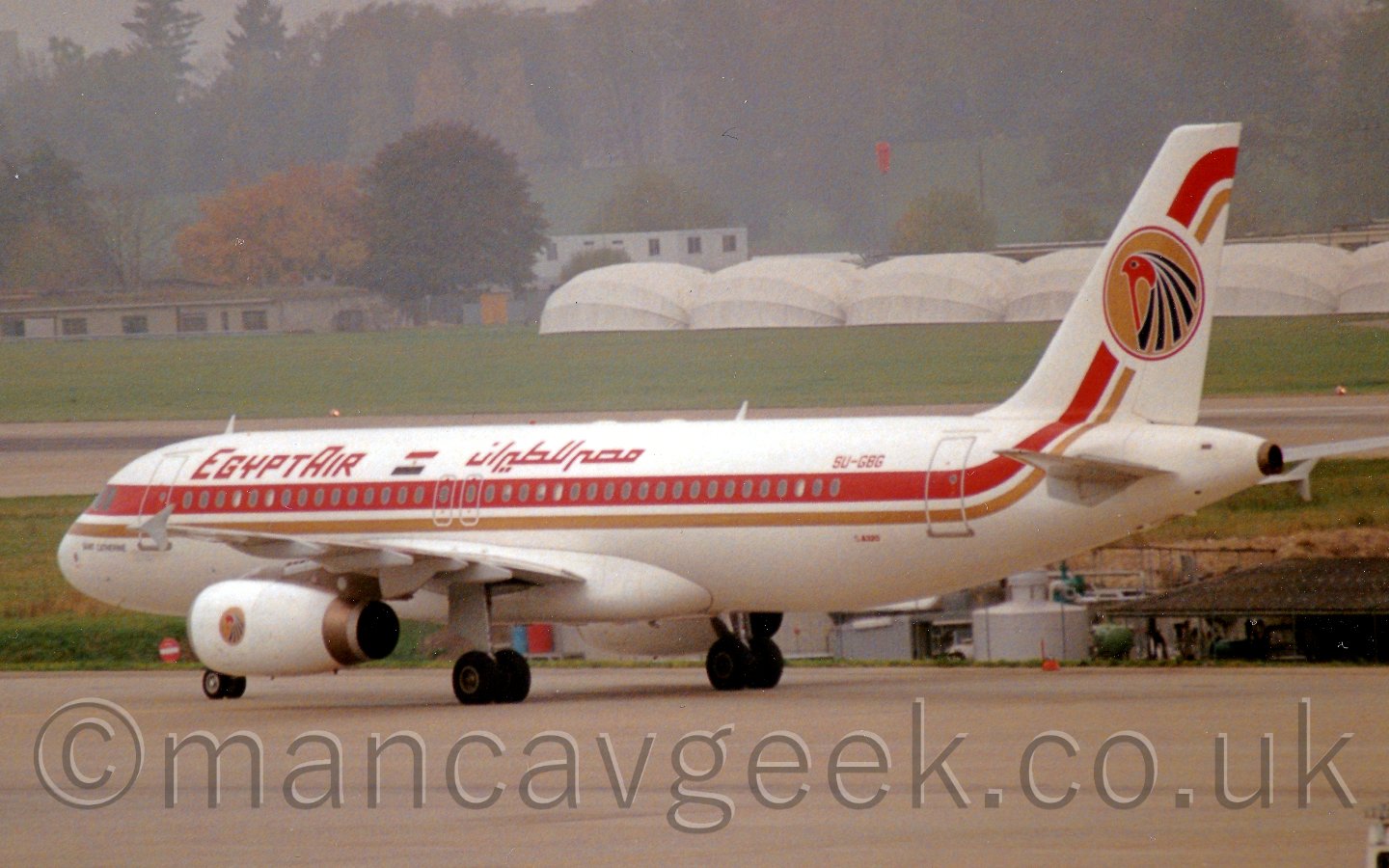 Side view of a twin engined jet airliner taxiing from right to left, facing slightly away from the camera. The plane is mostly white, with a thick red and thin golden stripe running along the body and up in to the tail. There are red EgyptAir titleson the upper forward fuselage, in English and Arabic, on either side of an Egyptian flag. On the tail, there is a golden circle outlined in red, bearing the head of a red bird with blue plumage, actually an image of Horus, one of the Ancient Egyptian Gods, a logo that also appears on the engine pods. In the background, large areas of grass lead off to what appears to be large white circular rigid tents in front of some low buildings, with a tree-covered hillside beyond that slowly vanishing in to haze.